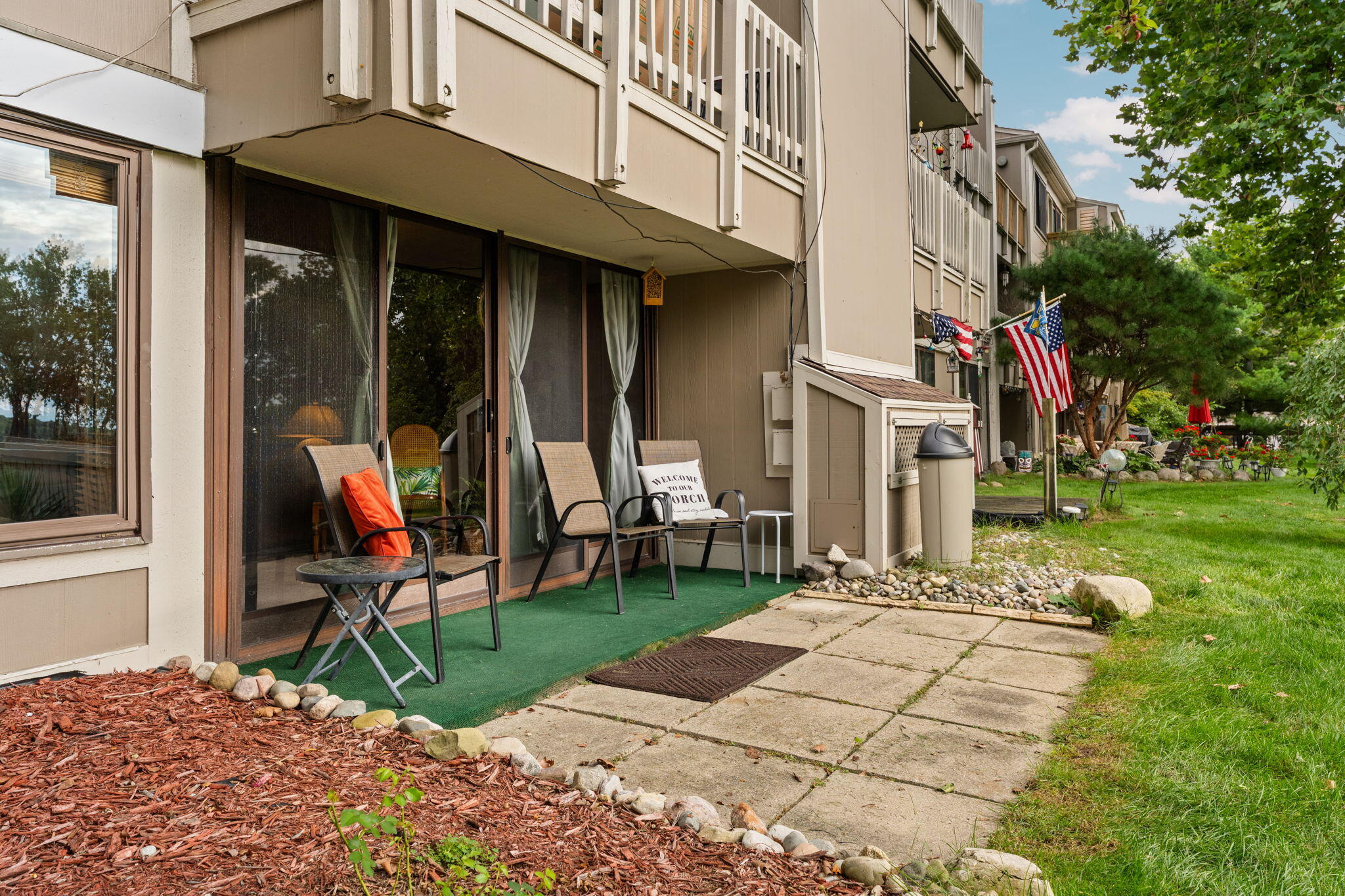Condo patio with chairs on a green rug beside sliding glass doors and a paved walkway.