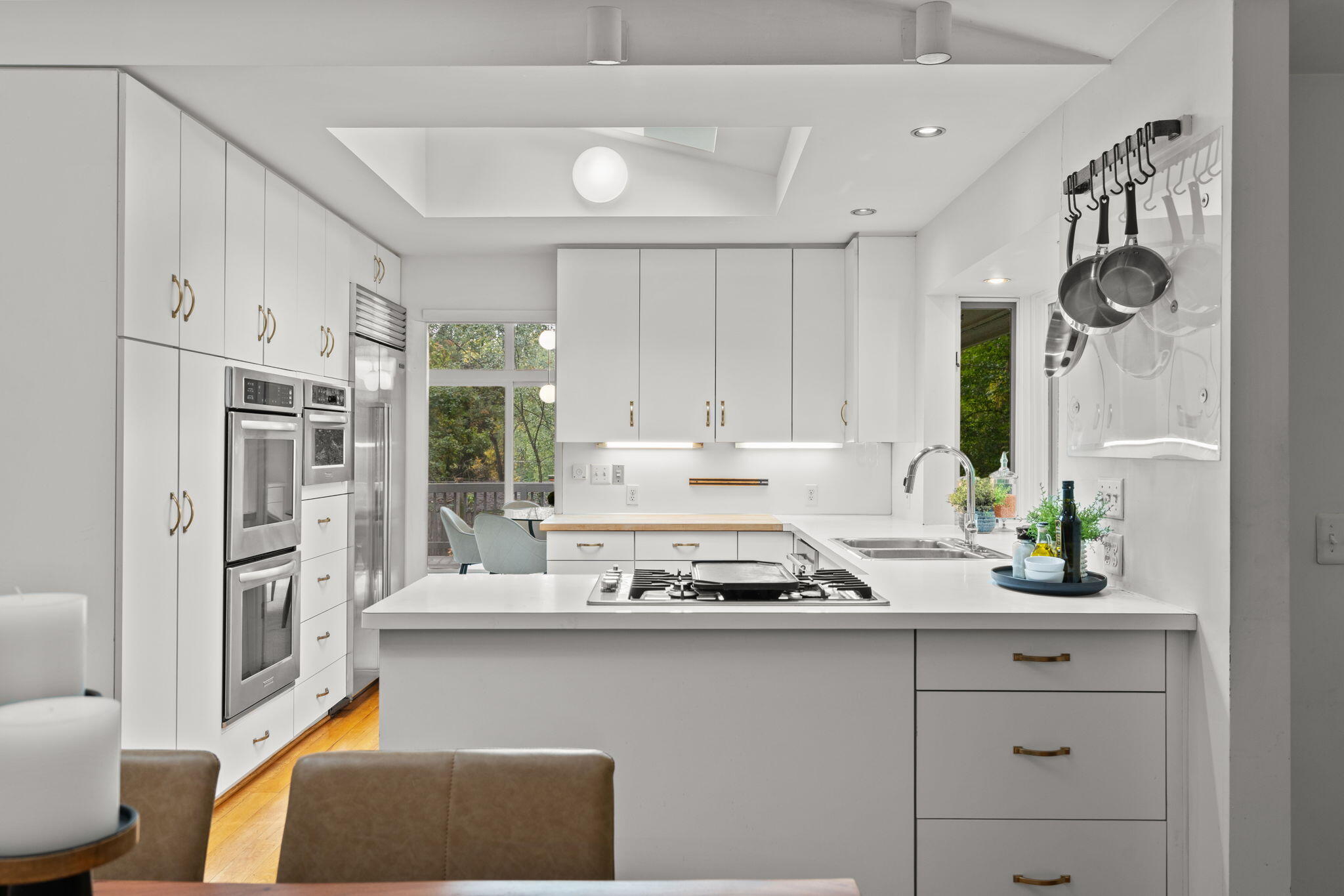 Bright white kitchen with double ovens, a central gas cooktop, and windows overlooking trees.