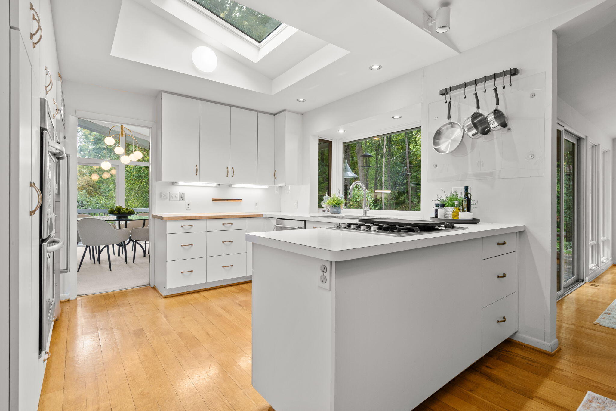 Bright white kitchen with a large cooktop island, skylight, and windows overlooking trees.