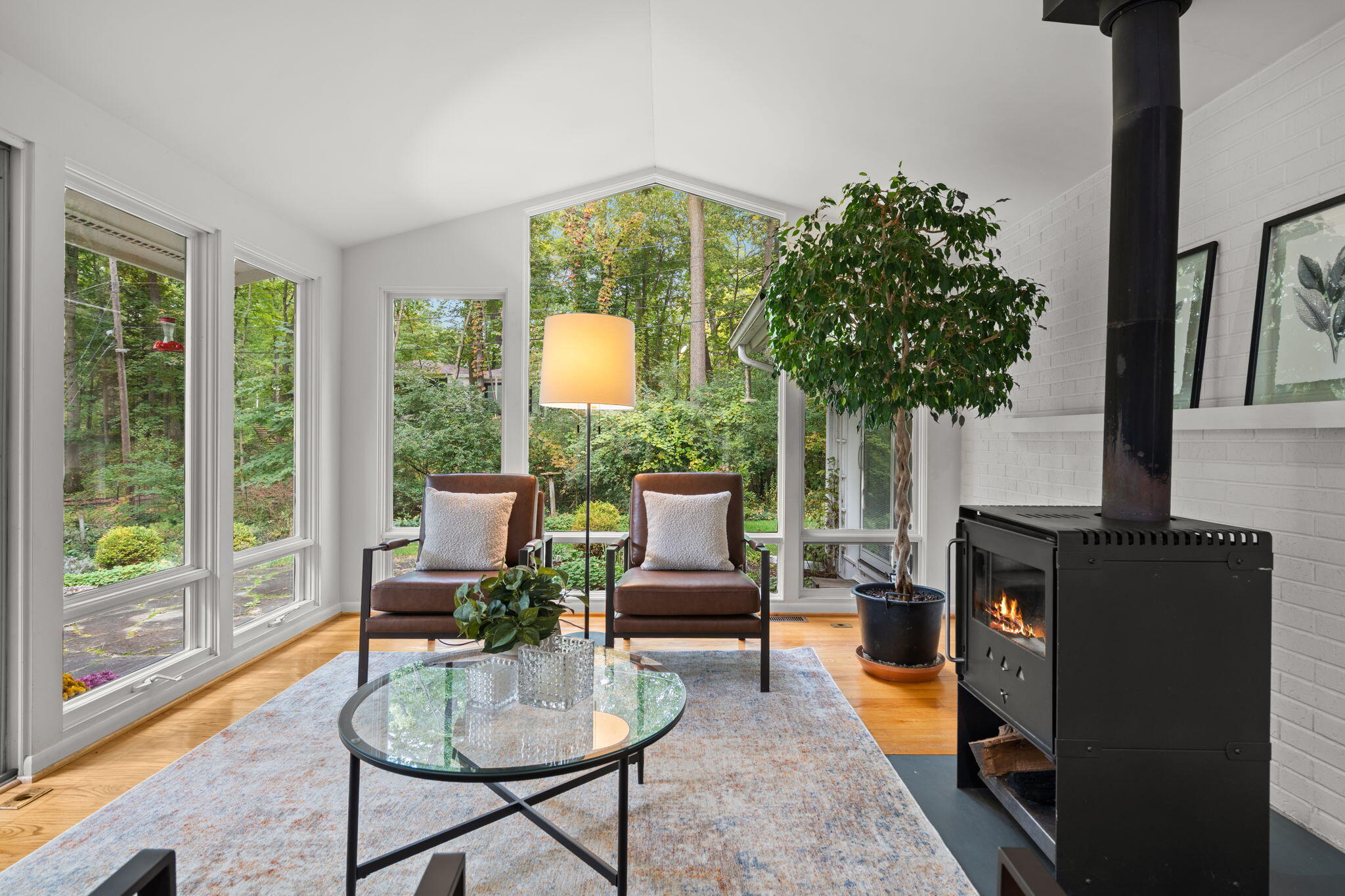 Sunroom with broad woodland-view windows, two leather chairs, and a lit wood-burning stove.