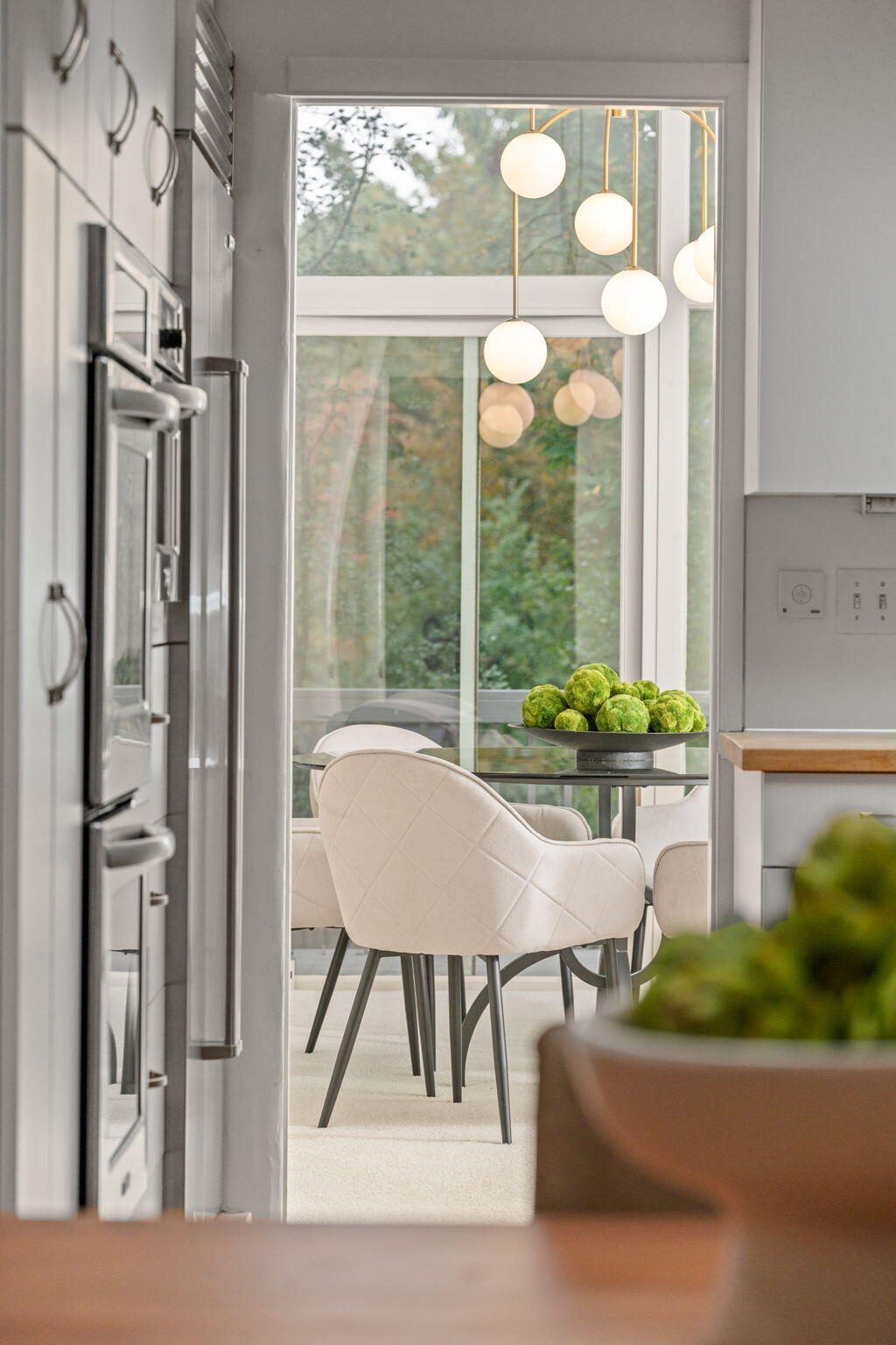 Dining area with white quilted chairs, globe pendant lights, and large windows overlooking trees.