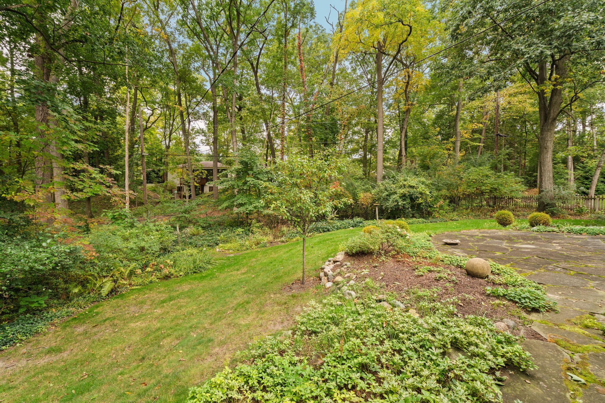 Wooded backyard with a stone patio, landscaped garden beds, and a house partly visible through the trees.