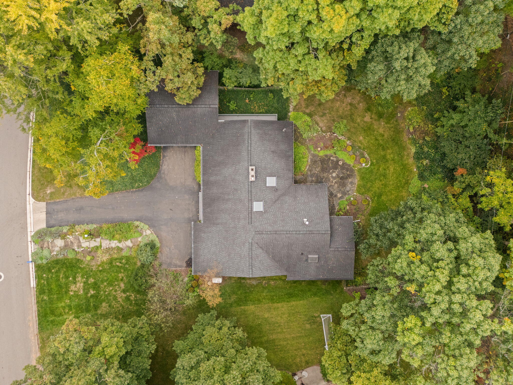 Aerial view of a house surrounded by mature trees, with a driveway, lawn, and stone patio.