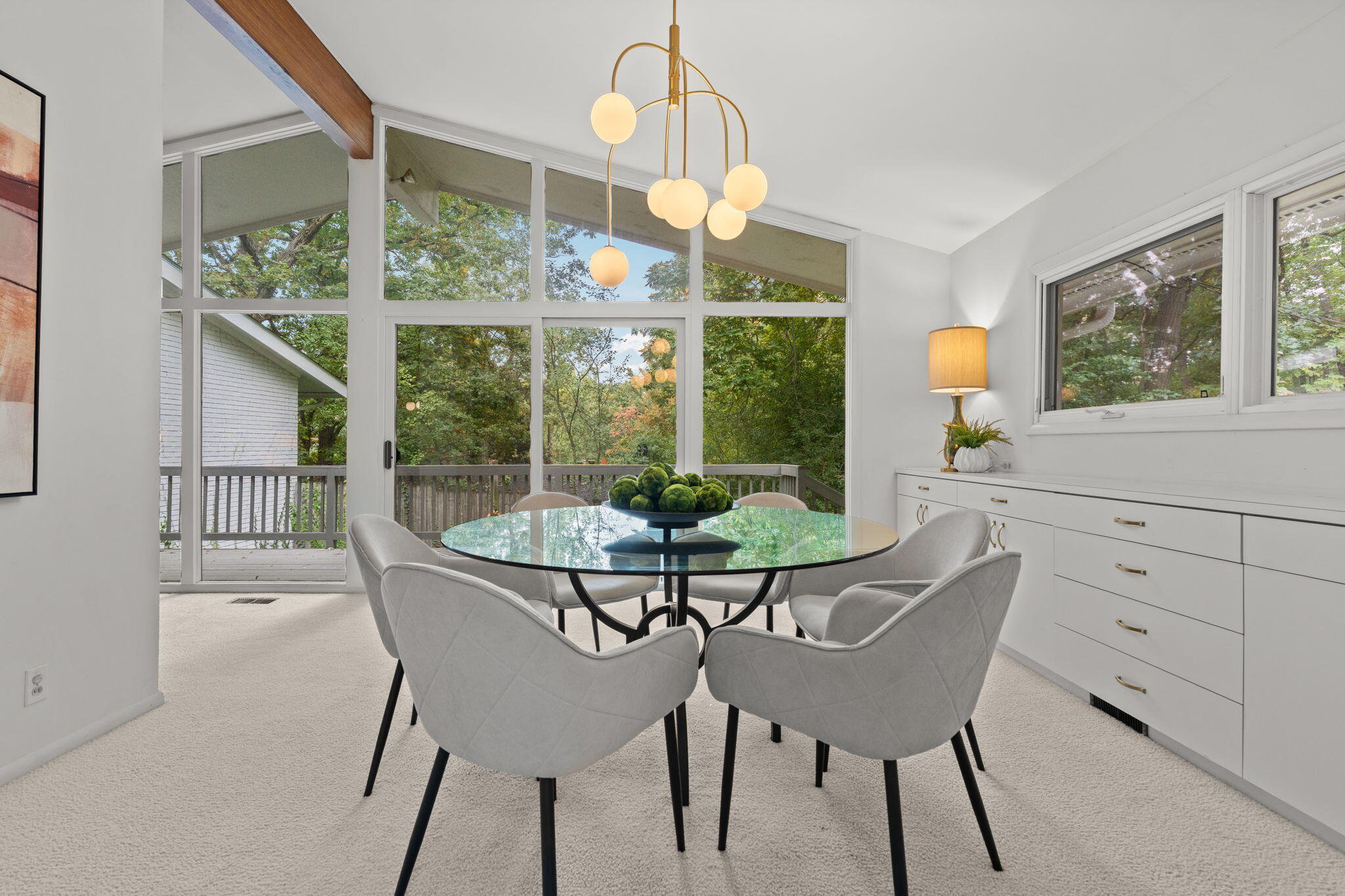 Dining room with a glass table, upholstered chairs, and large windows overlooking a wooded deck.