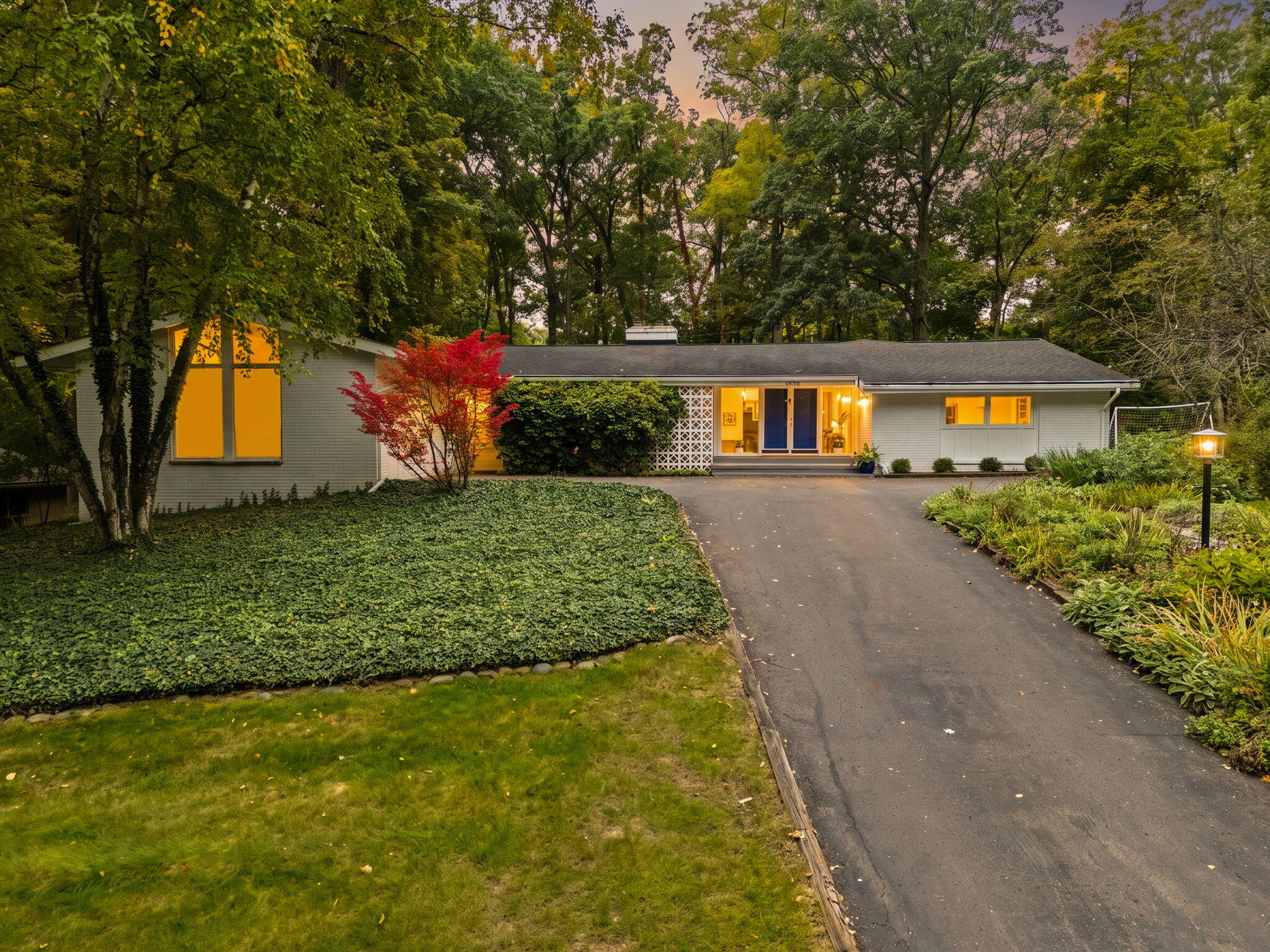 Single-story home with a long driveway, landscaped yard, and warmly lit windows at dusk.