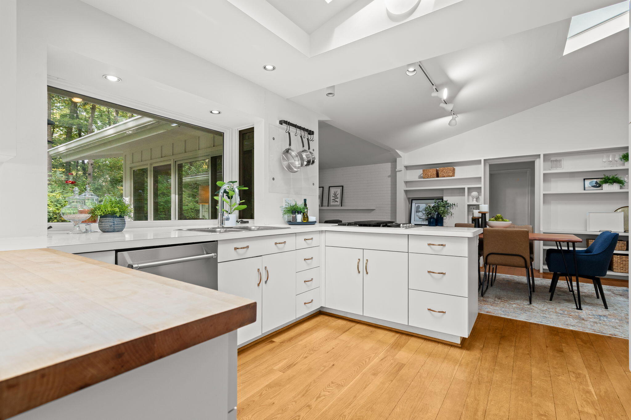 Bright white kitchen with a large island, vaulted ceiling, wood floors, and a window overlooking trees.