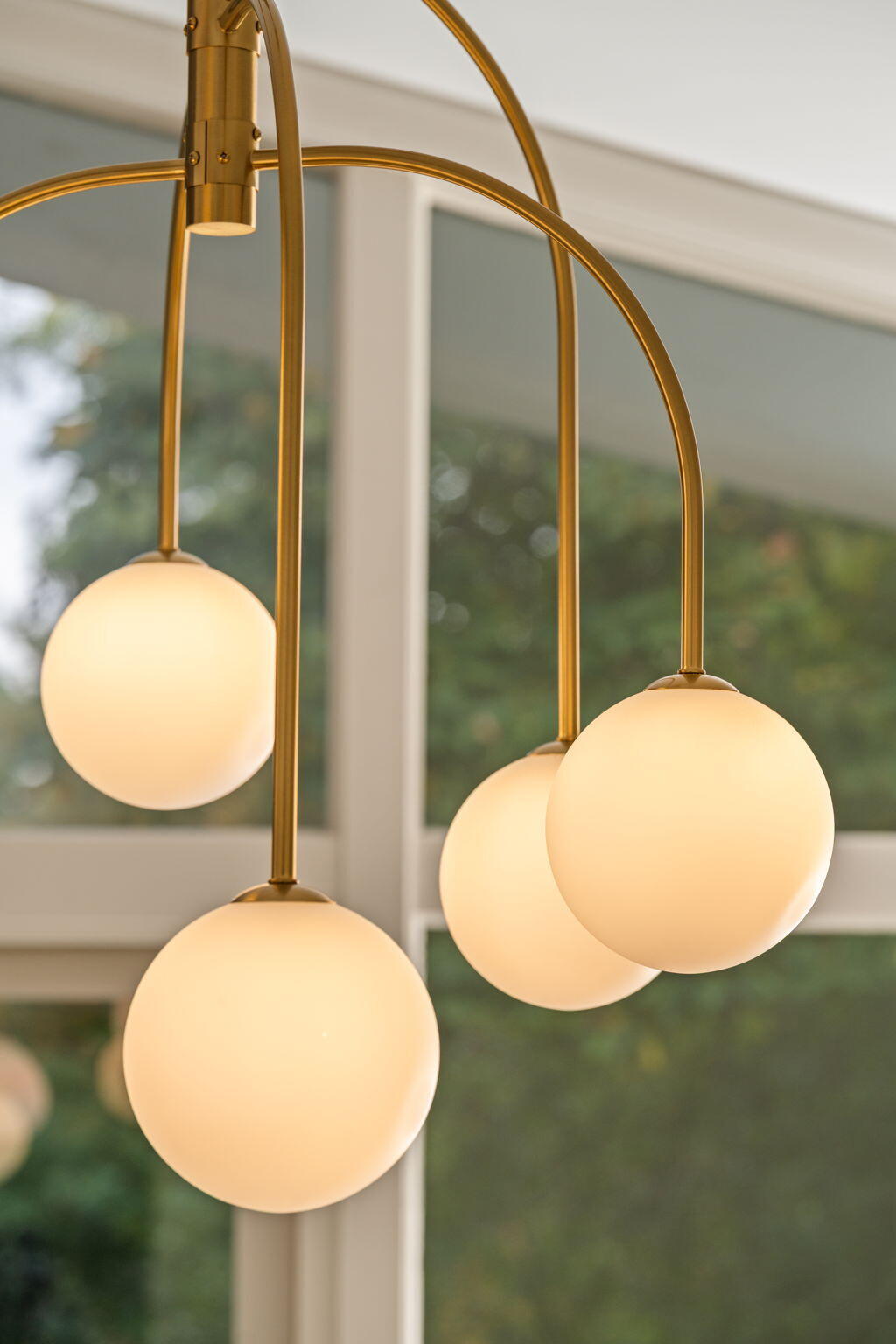 Brass chandelier with glowing spherical shades in front of tall windows.