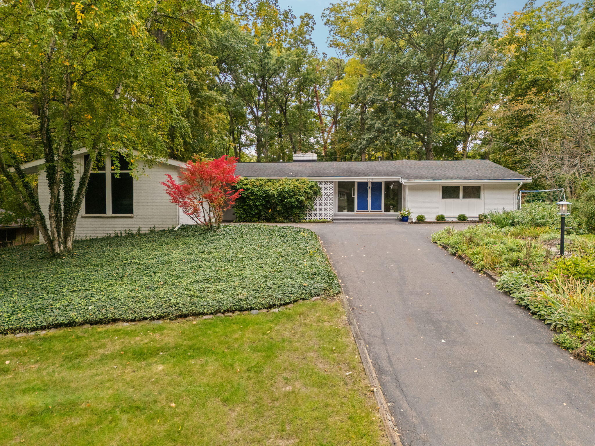 Single-story white brick home with blue double doors, a broad driveway, and mature autumn landscaping.