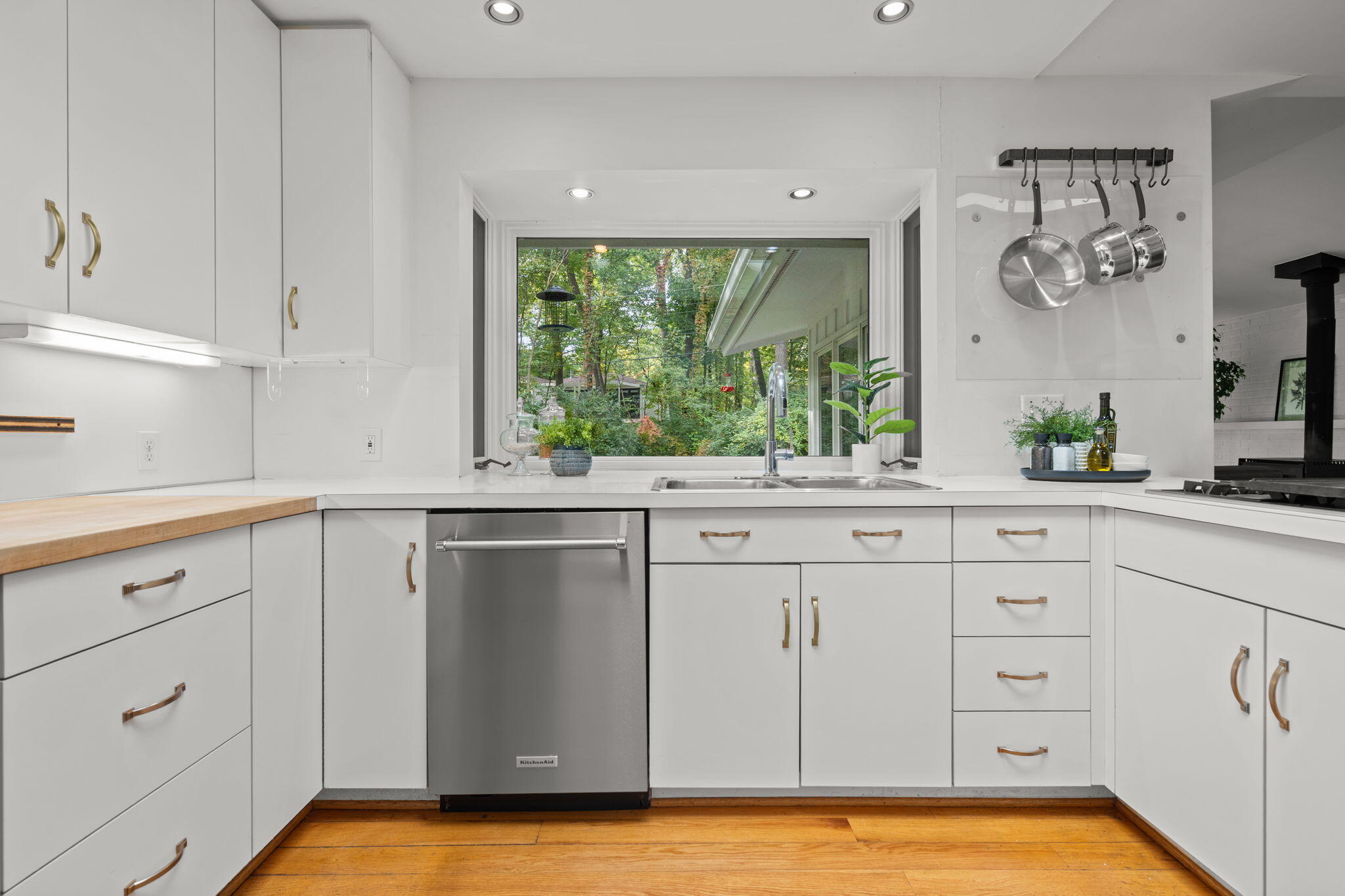 White kitchen with brass cabinet pulls, a stainless dishwasher, and a sink overlooking leafy trees.