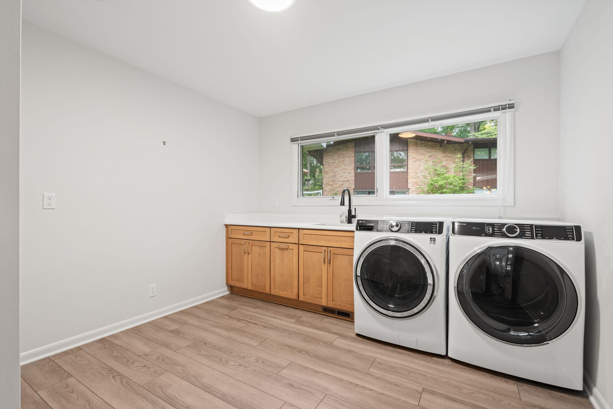 Laundry room with front-loading washer and dryer, wood cabinets, and a wide window above the sink.