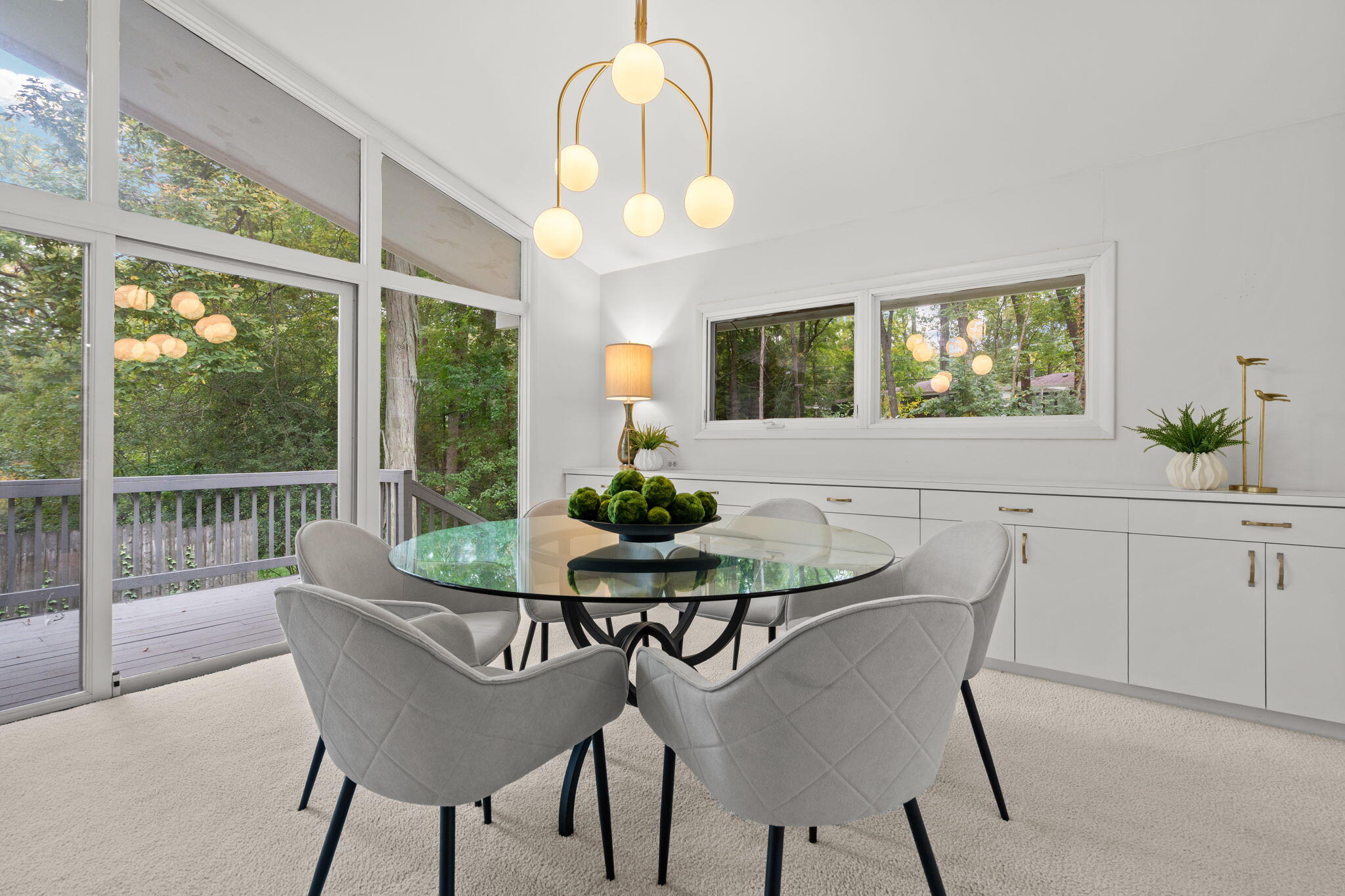 Dining room with a glass table and gray chairs beside large windows overlooking trees.