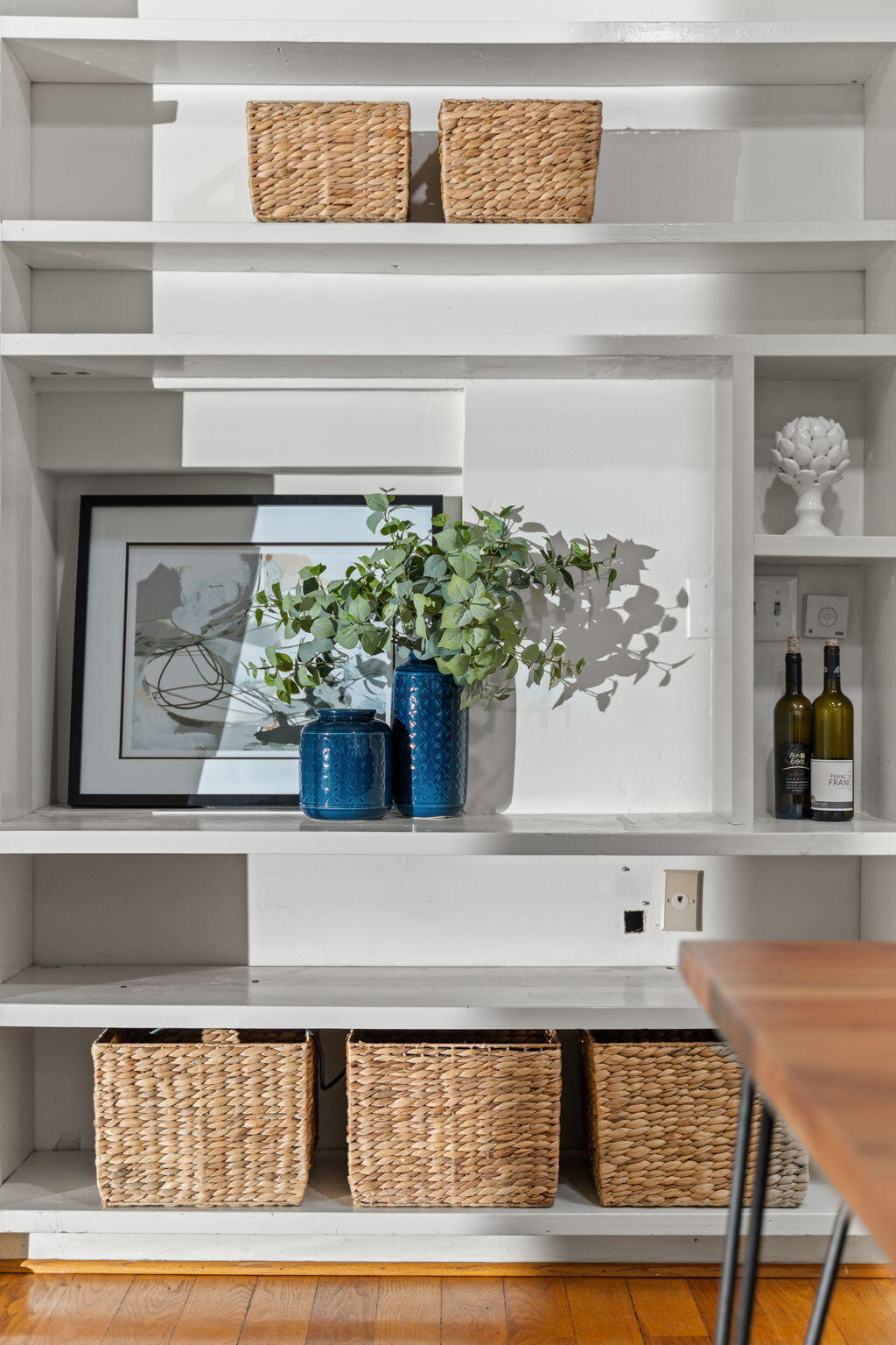 White built-in shelves with woven baskets, blue vases, framed art, and wine bottles.