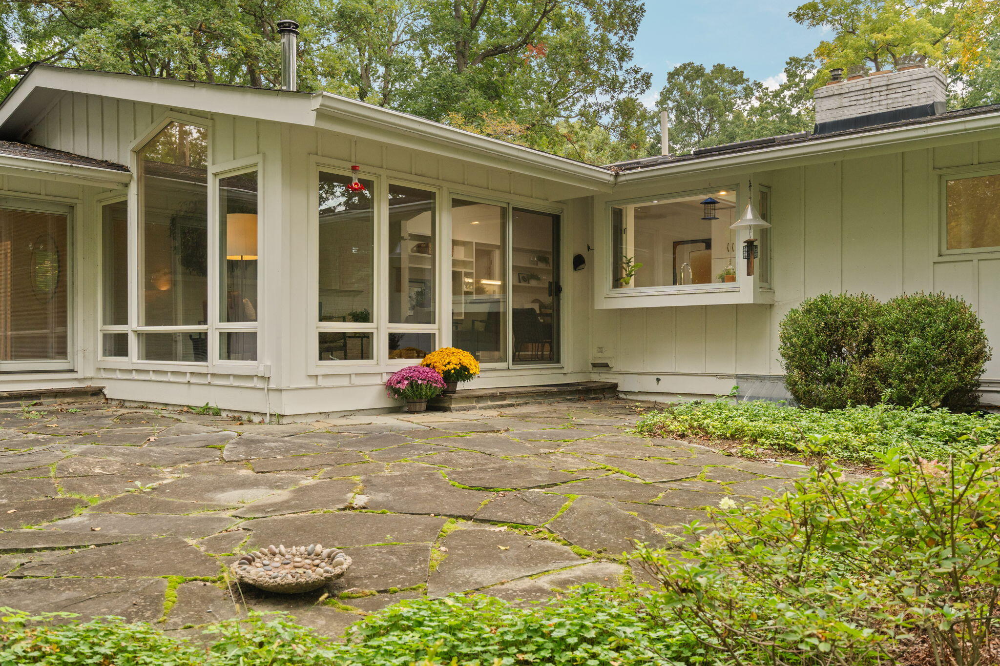Home’s rear patio with expansive windows, flagstone paving, and potted chrysanthemums.