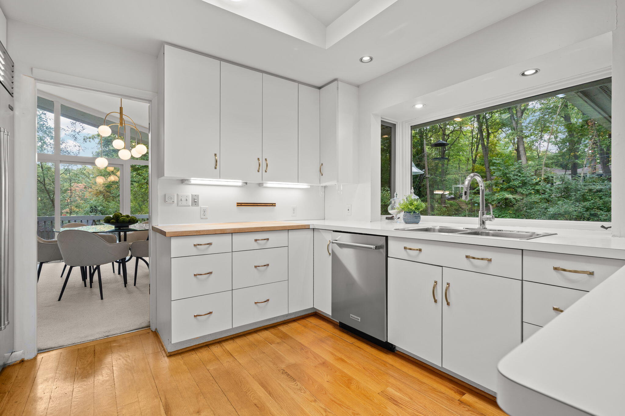 White kitchen with brass pulls, hardwood floors, and a large window overlooking leafy trees.