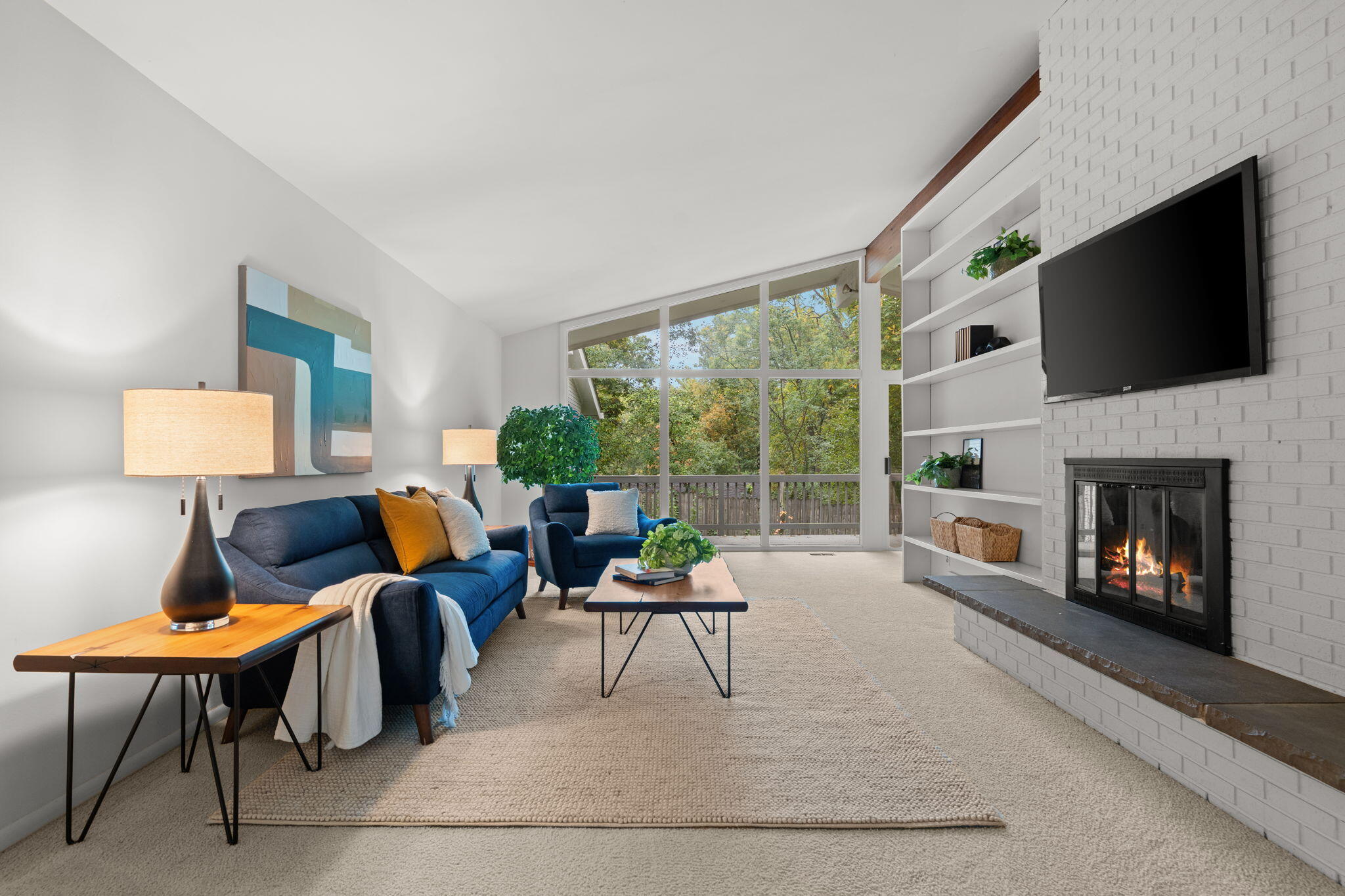 Staged living room with blue sofas, a white brick fireplace, and large windows overlooking trees.