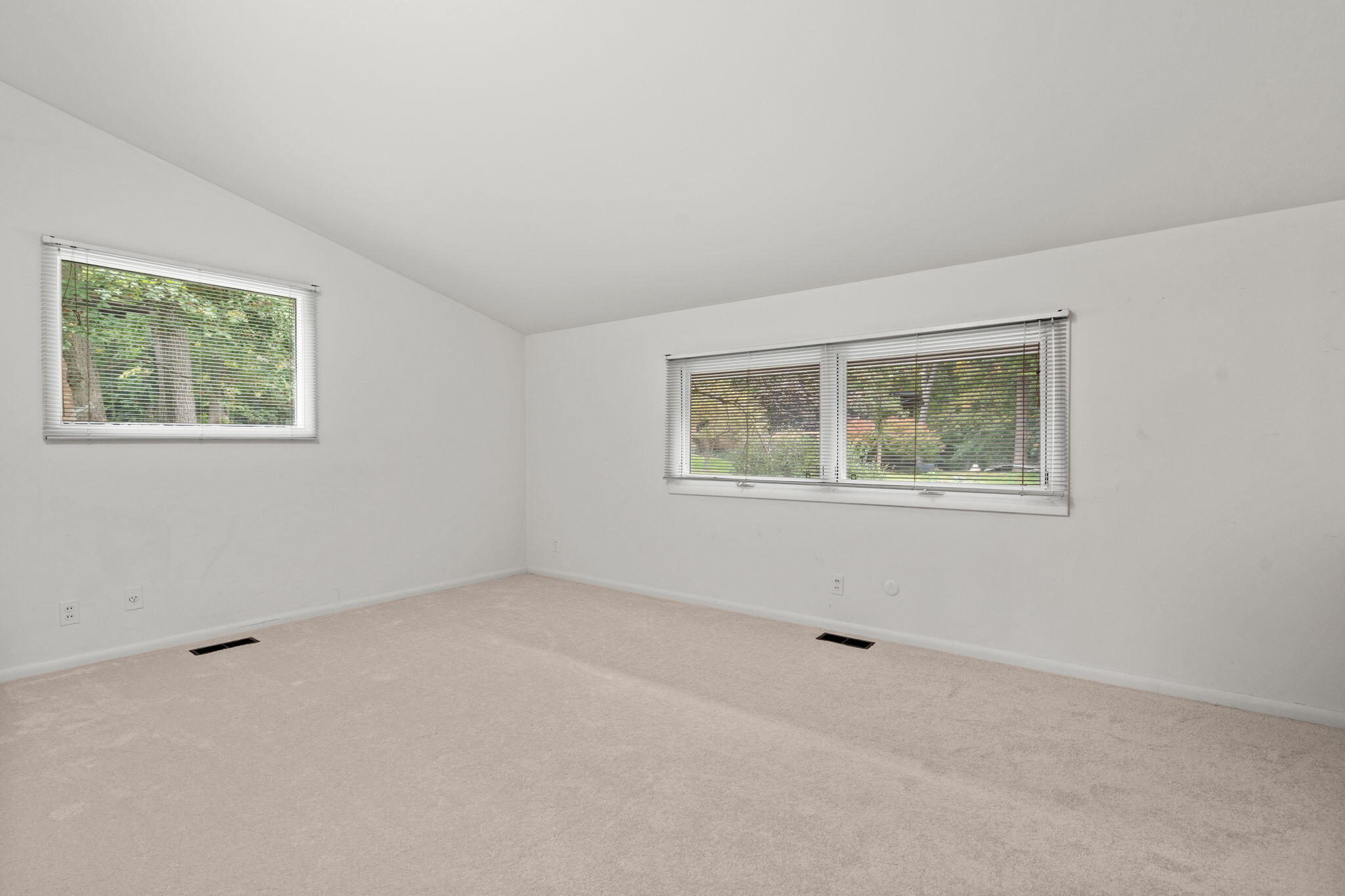 Empty carpeted room with two windows overlooking greenery.
