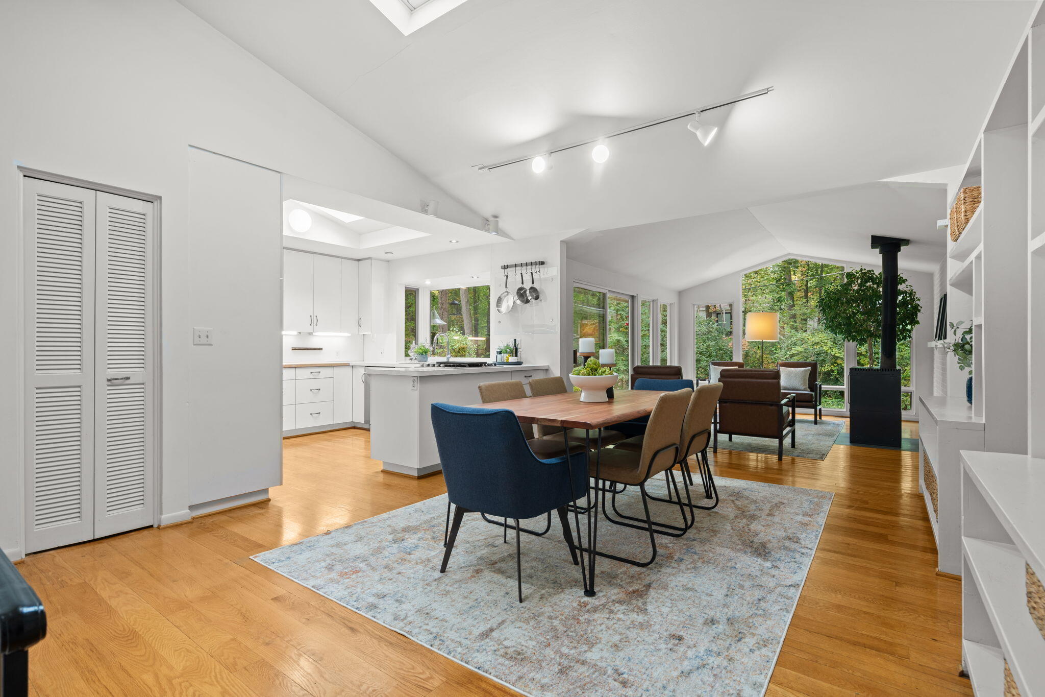 Open dining area with a wood table and blue chair, adjacent to a bright white kitchen and windowed living room.