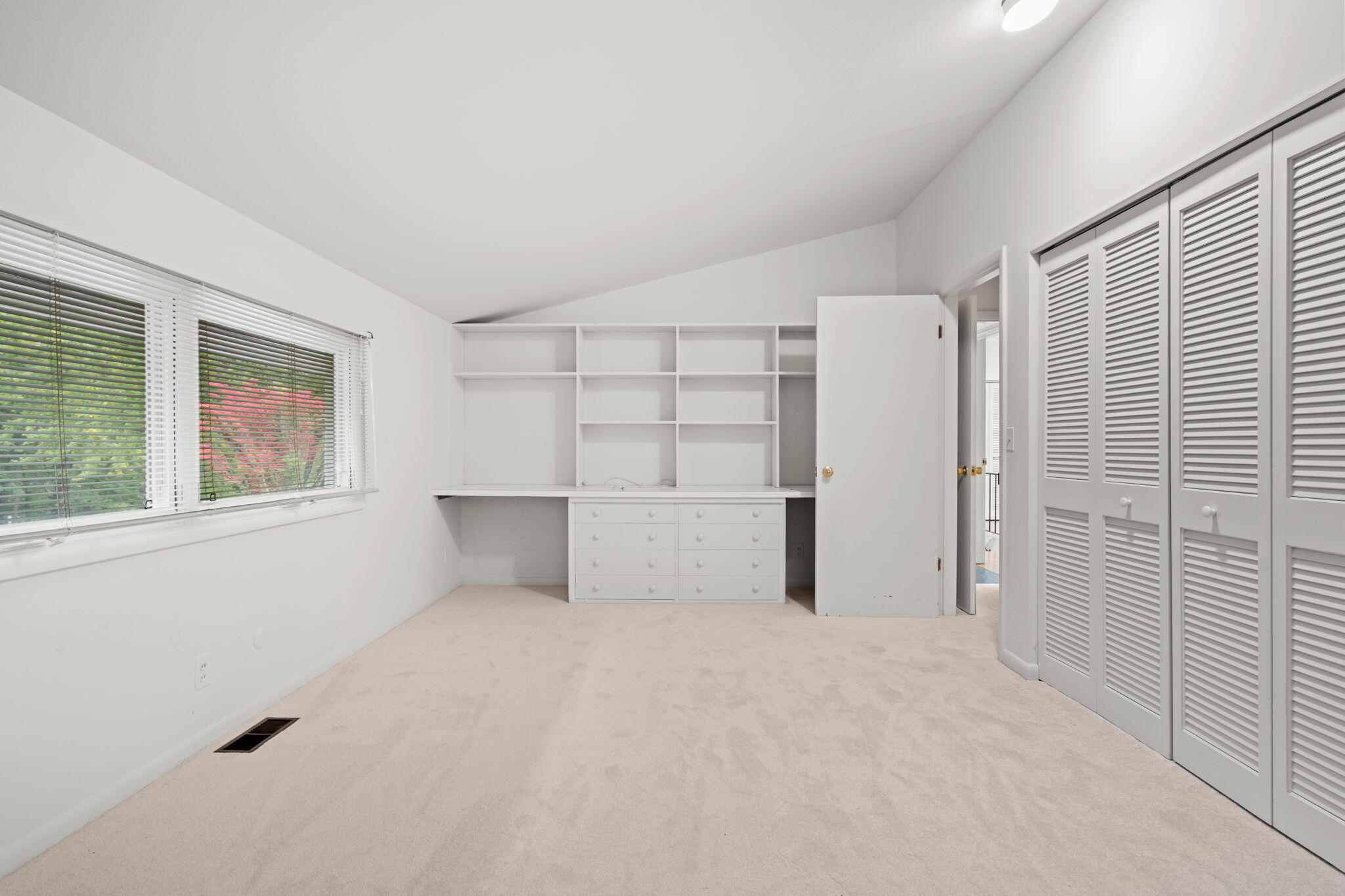 Empty carpeted room with built-in shelving and desk, louvered closets, and windows with blinds.