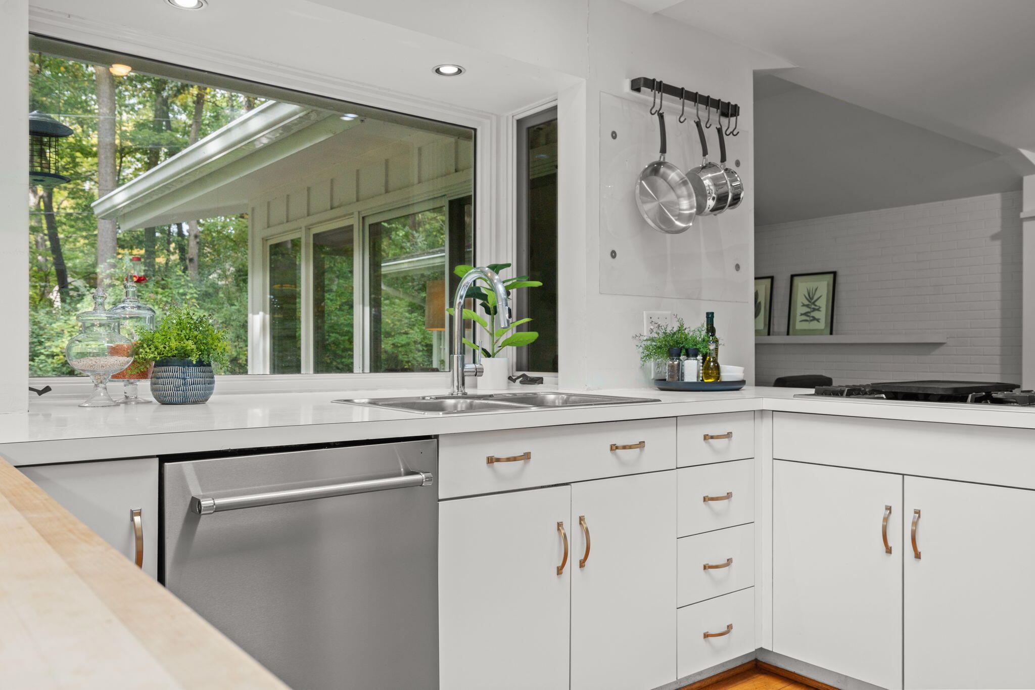 Bright white kitchen with a stainless dishwasher, hanging cookware, and large windows overlooking trees.