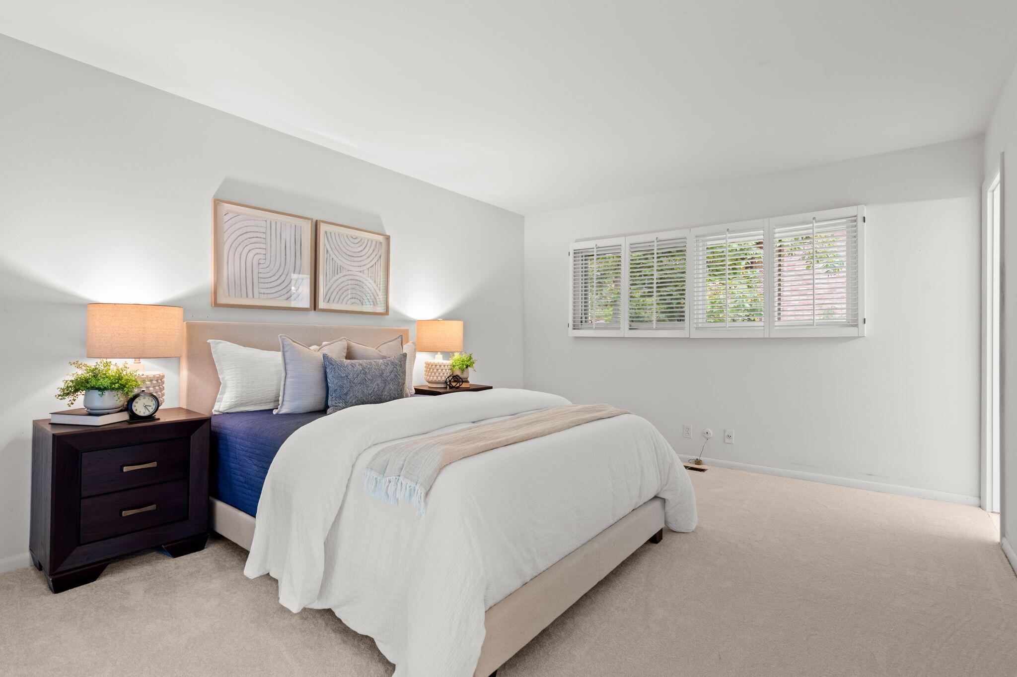 Staged bedroom with a white-and-blue bed, matching bedside lamps, and a wide shuttered window.