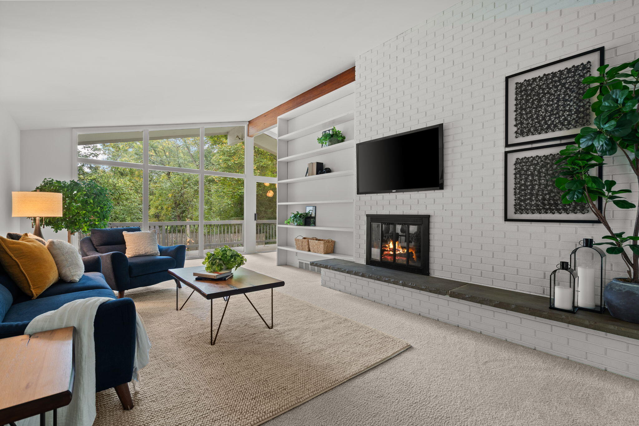 Living room with a white brick fireplace, blue seating, and large windows overlooking trees.