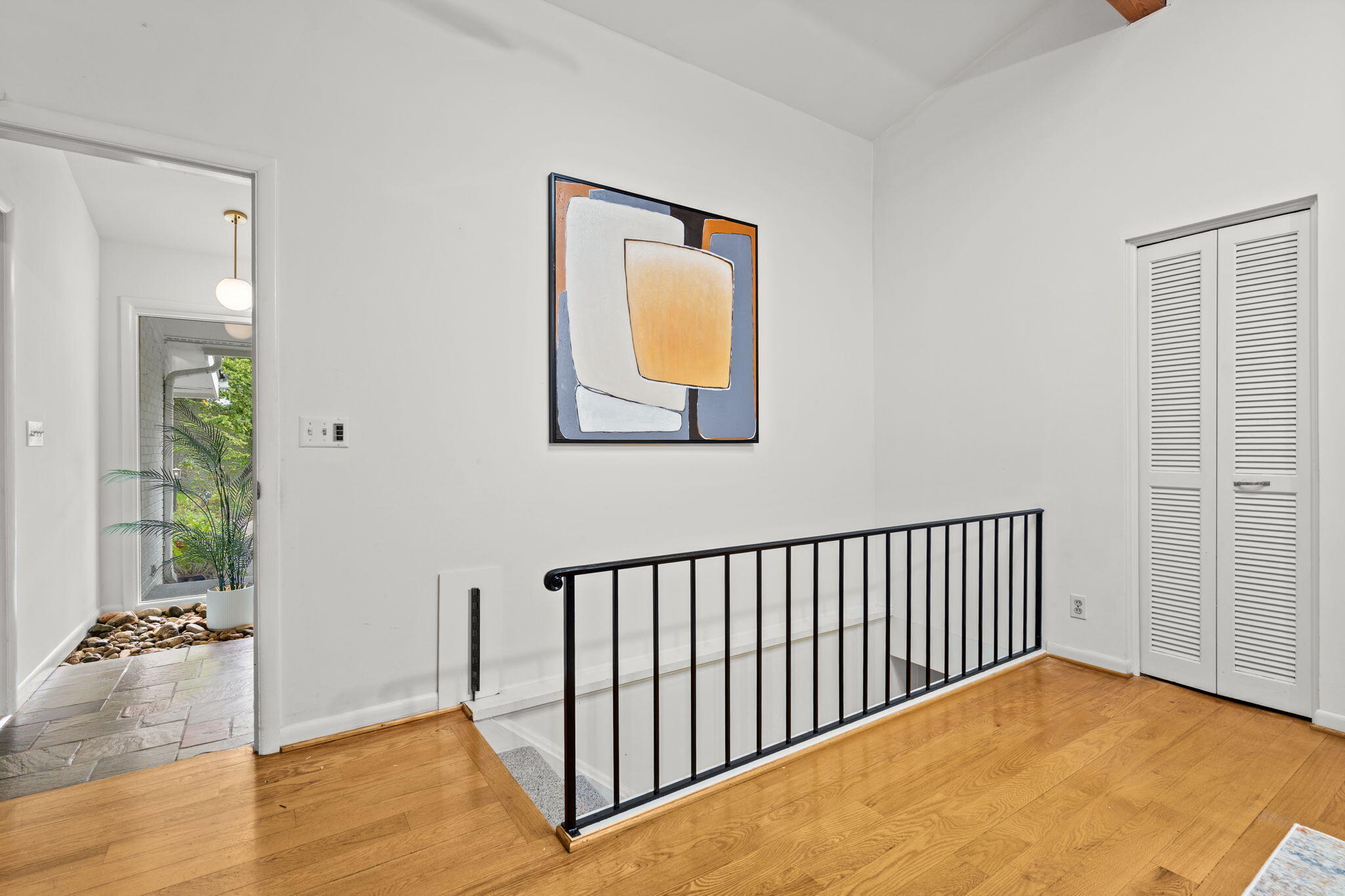 Bright upstairs landing with hardwood floors, black stair railing, abstract artwork, and louvered closet doors.