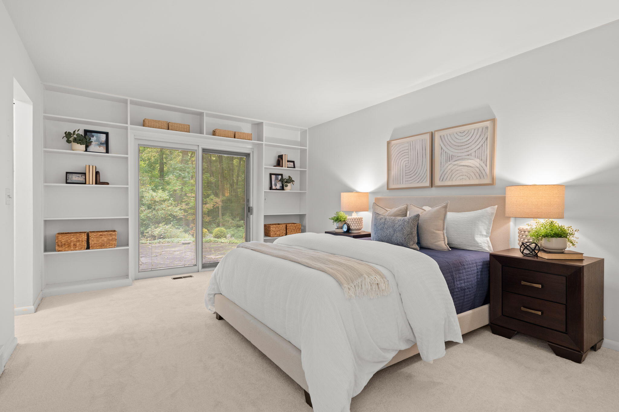 Staged bedroom with built-in shelving, a large bed, and sliding glass doors facing leafy trees.