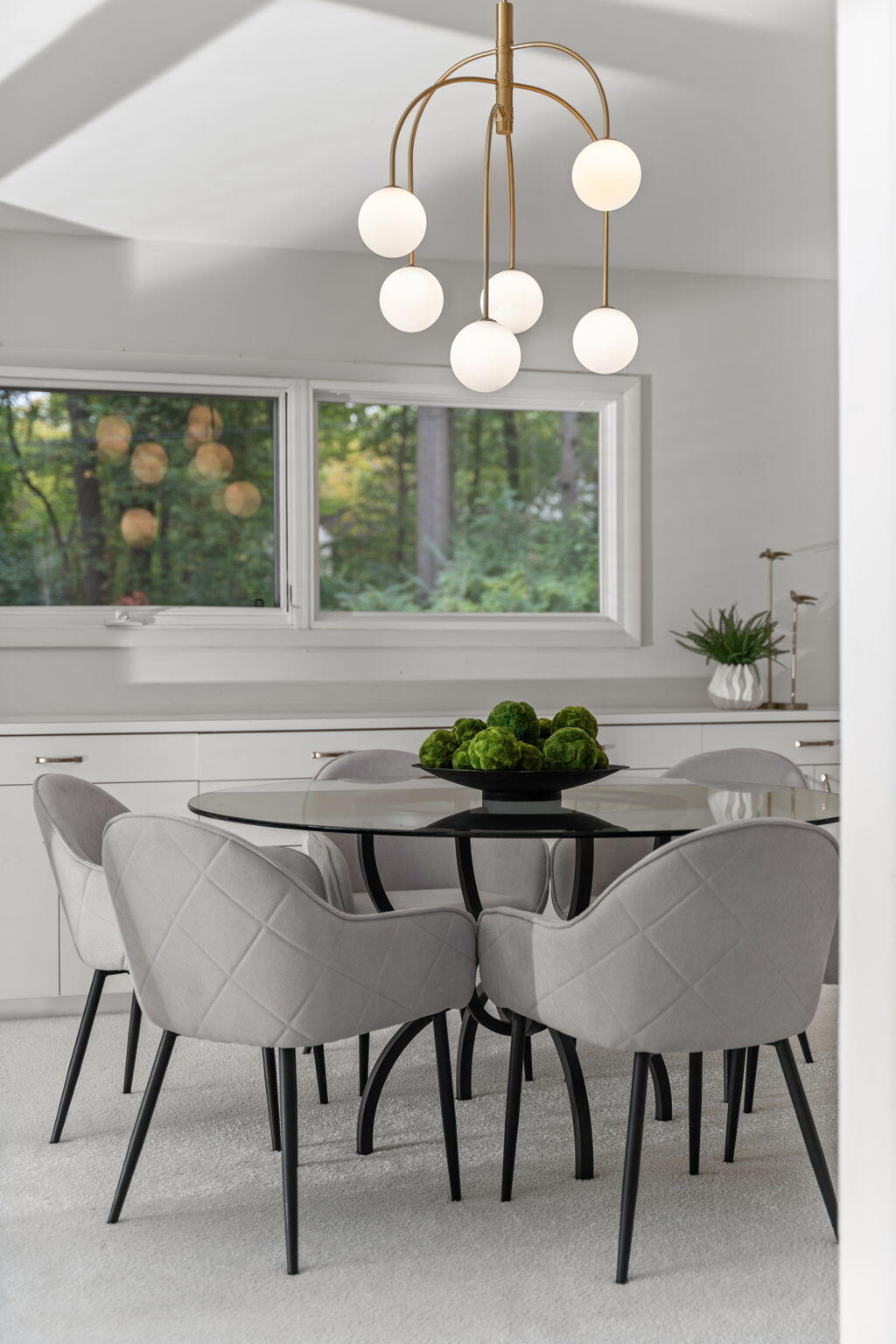 Dining area with a glass table, gray chairs, and a brass globe chandelier by large windows.