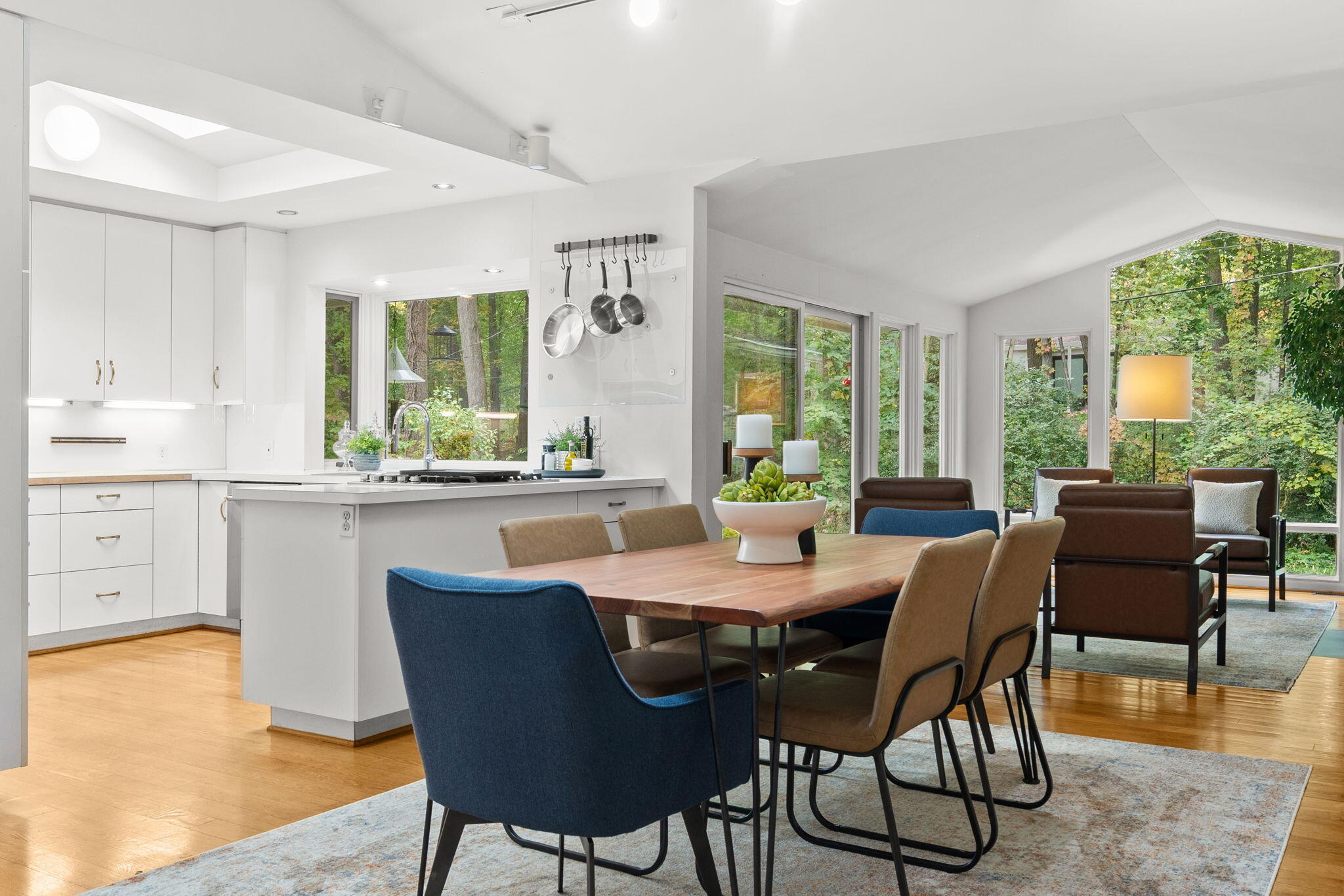 Open-plan kitchen and dining area with white cabinetry, a wooden table, and large windows facing trees.