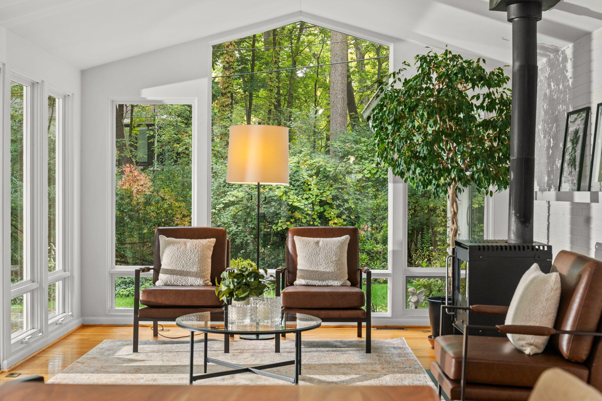 Bright living room with tall wooded-view windows, brown leather chairs, and a black wood stove.