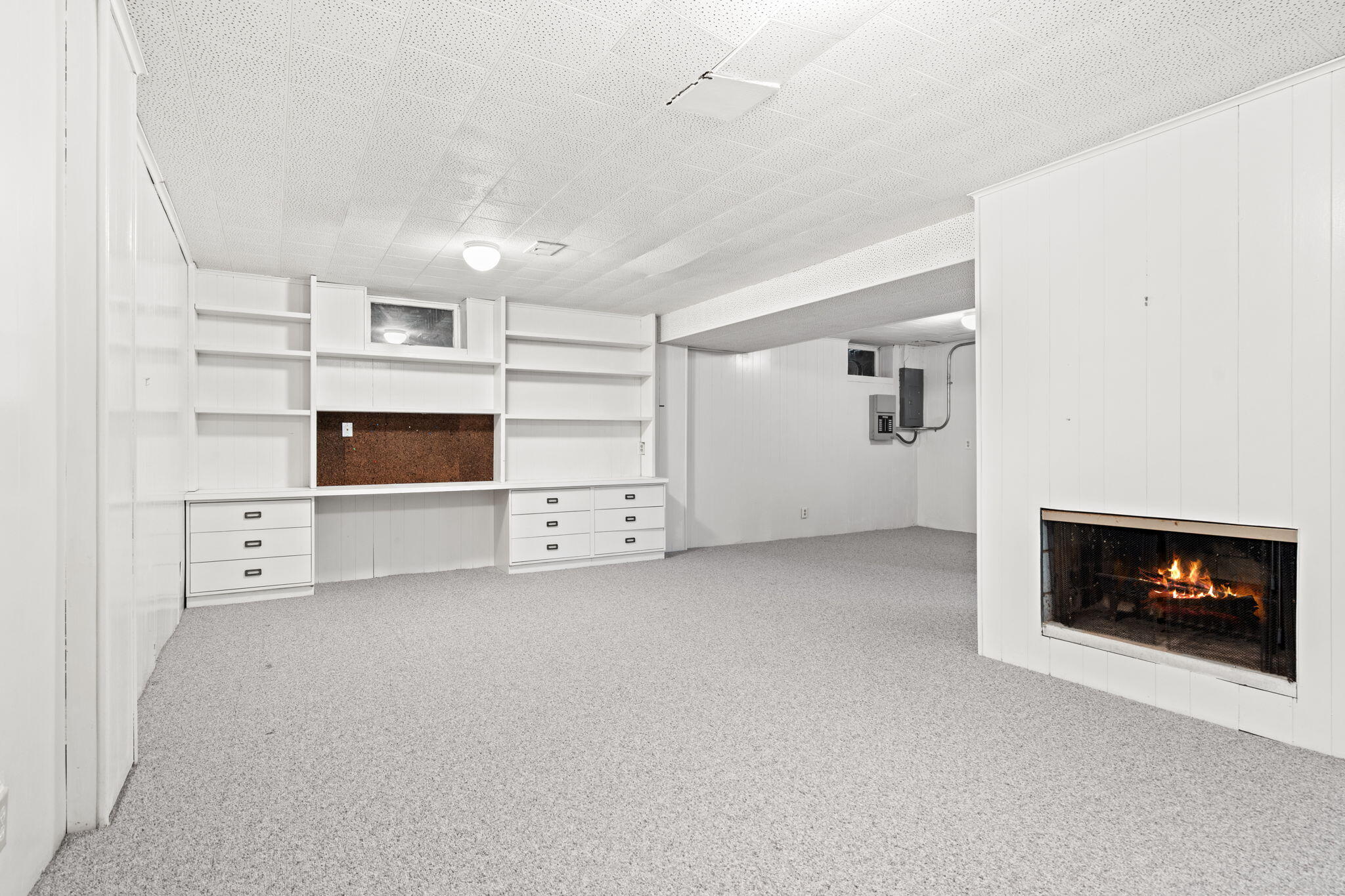 Basement room with built-in shelving and drawers, gray carpet, and a lit fireplace.