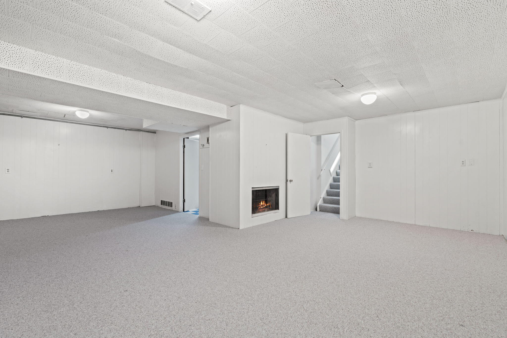 Vacant carpeted basement room with a lit fireplace and staircase.
