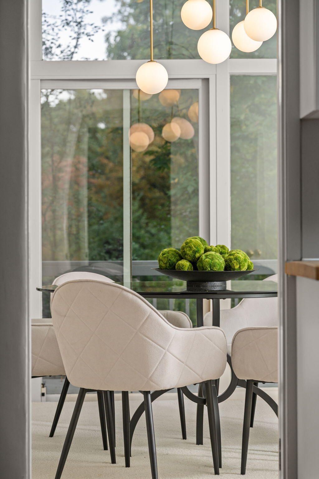 Dining area with cream chairs, a black table, globe pendants, and large windows overlooking greenery.