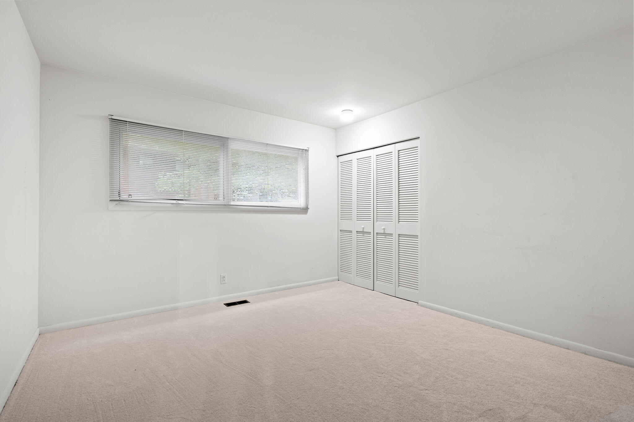 Empty bedroom with carpet, a wide window with blinds, and louvered closet doors.