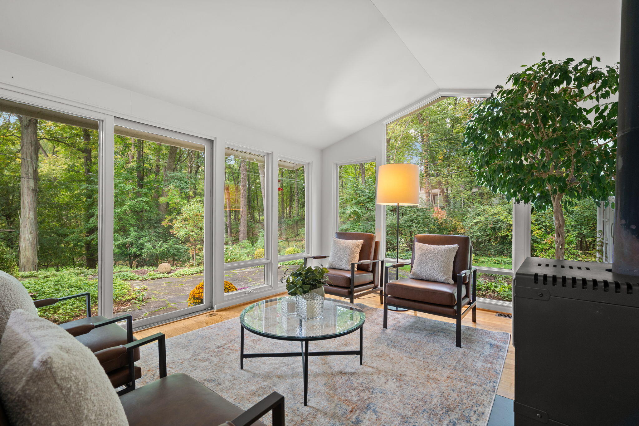 Living room with broad windows overlooking woods, brown armchairs, and a black stove.