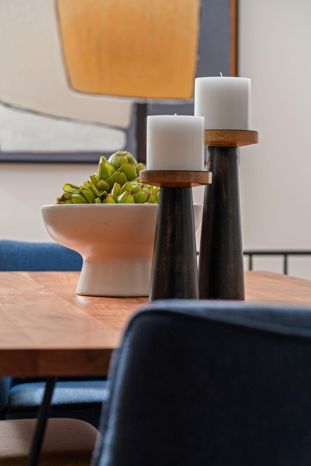 Dining table styled with white candles and a bowl of artichokes, surrounded by blue upholstered chairs.