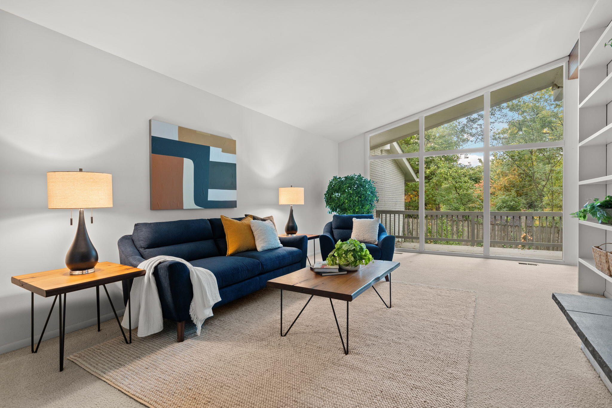 Staged living room with navy seating, abstract art, and large windows overlooking trees and a deck.