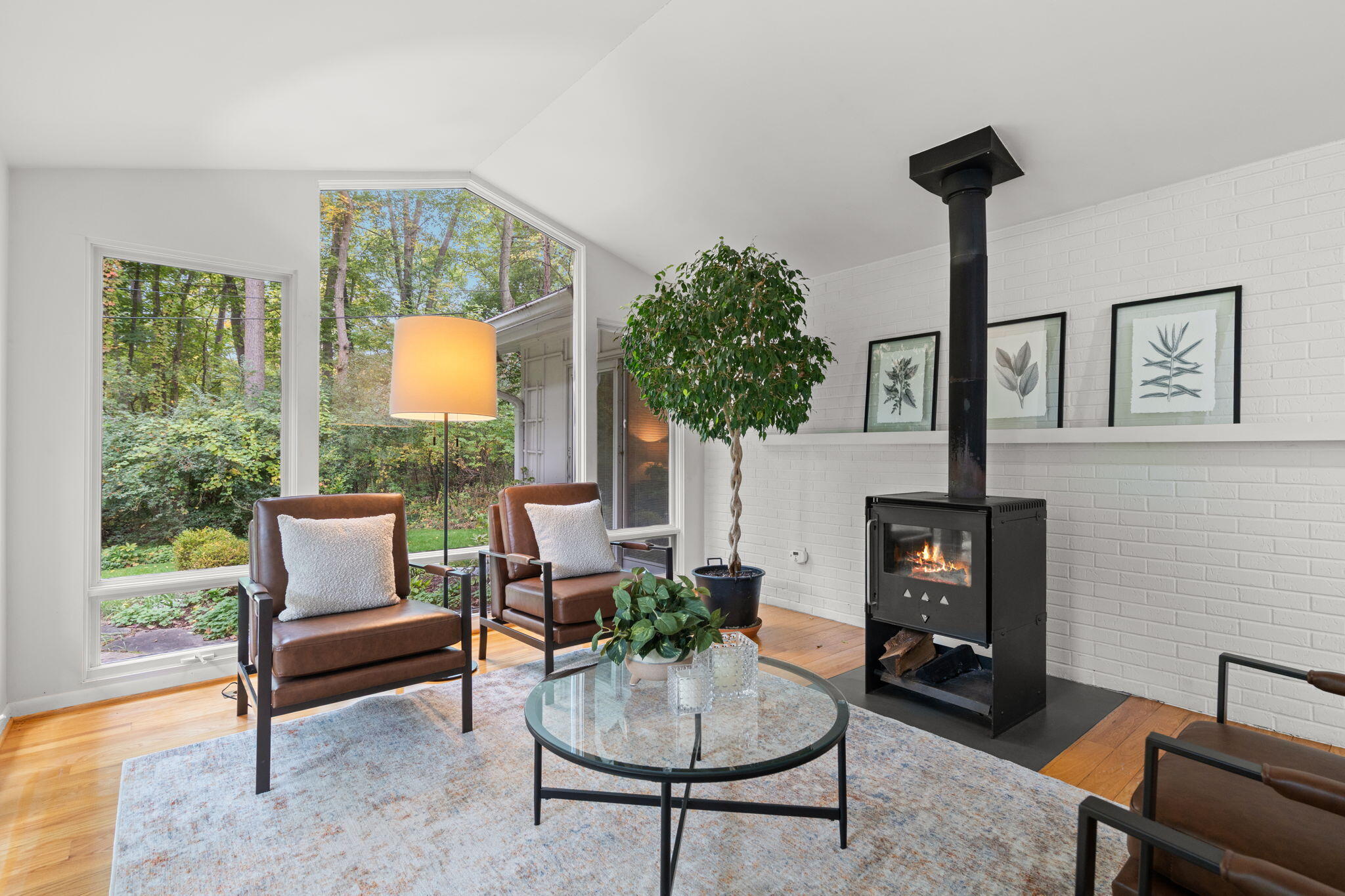 Living room with a black wood stove, brown leather chairs, and large windows overlooking trees.