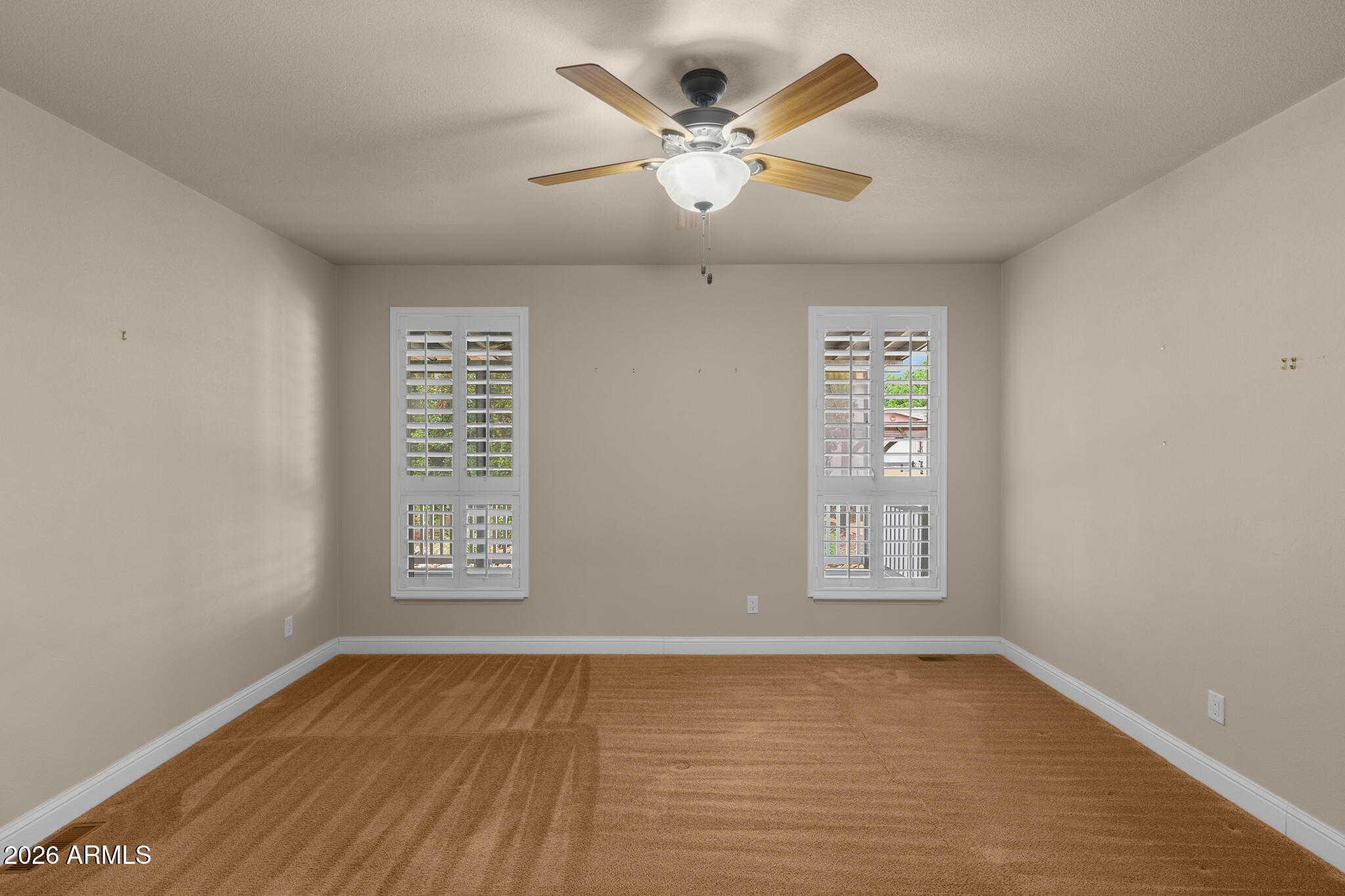 Empty carpeted room with two shuttered windows and a wood-blade ceiling fan.