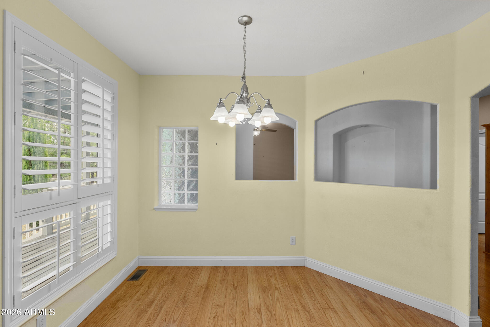 Empty room with hardwood floors, plantation shutters, a glass-block window, and a chandelier.
