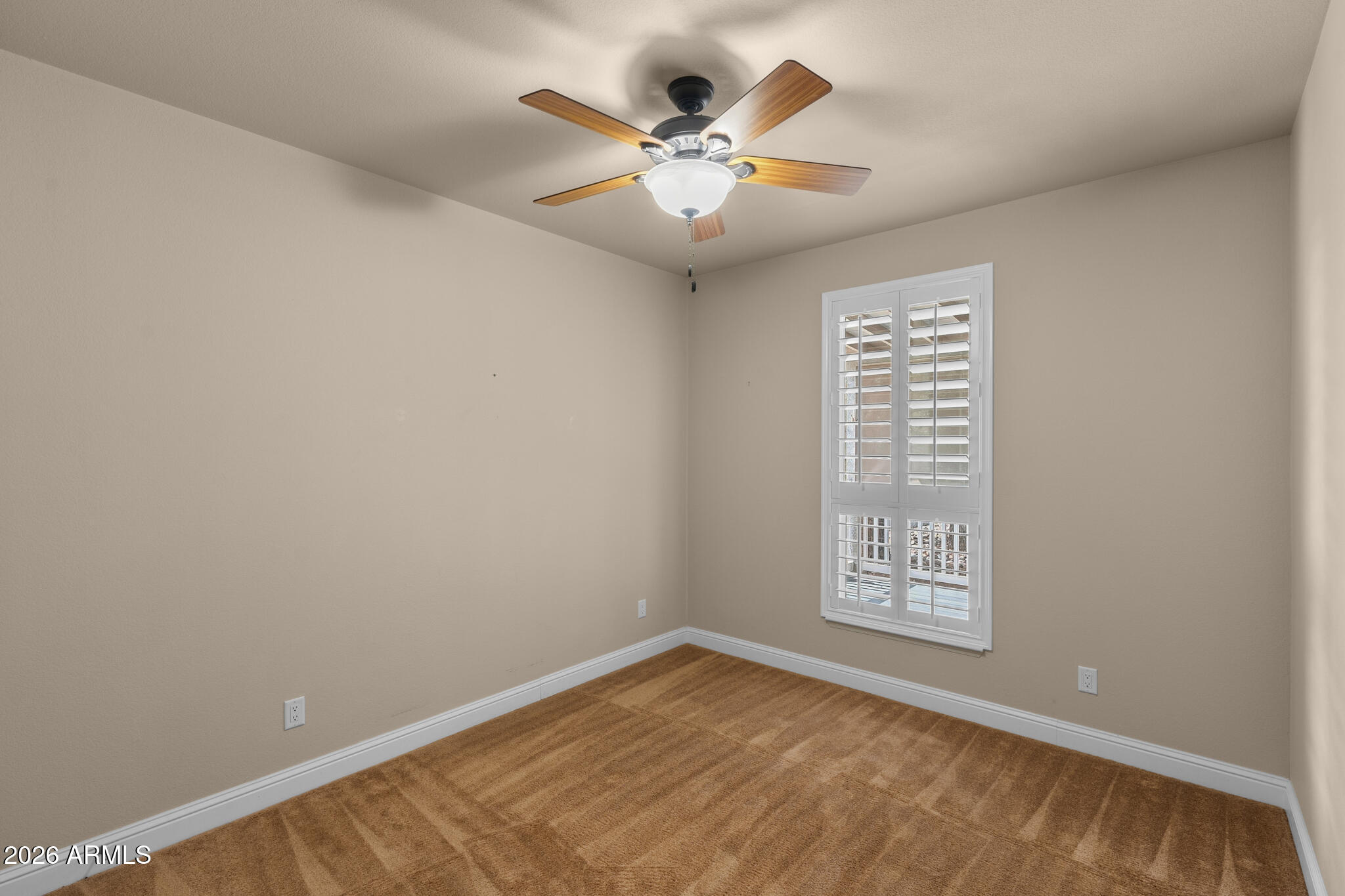 Empty bedroom with brown carpet, a wood-blade ceiling fan, and white plantation shutters.