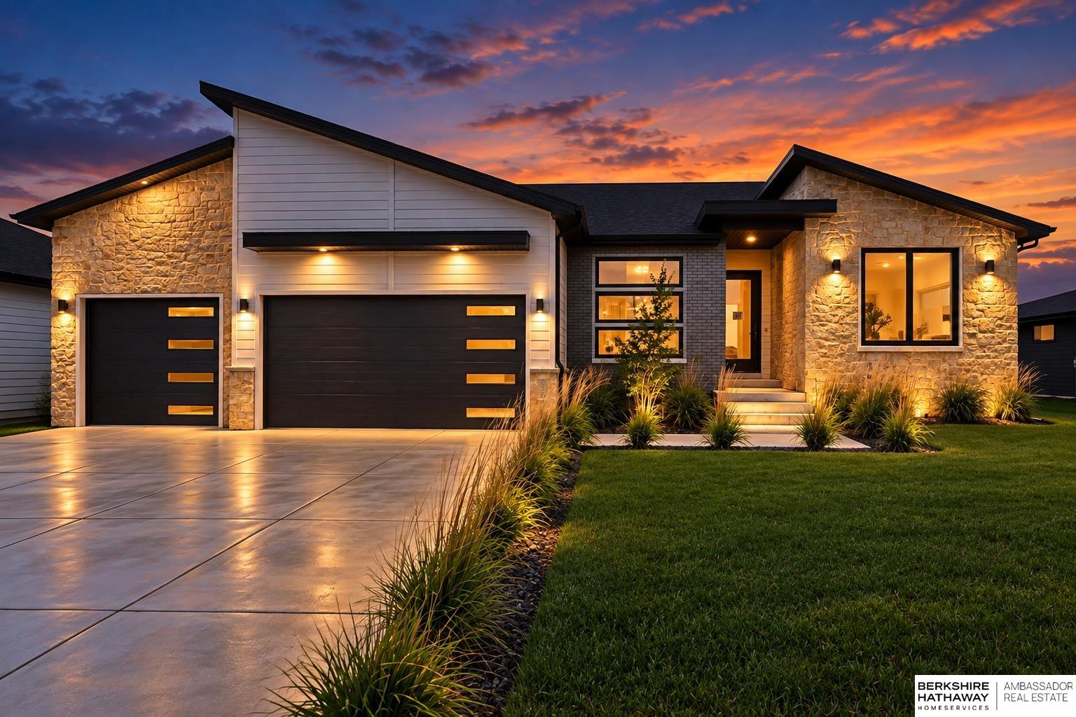 Modern stone-and-siding home at dusk with dark garage doors and illuminated landscaping.