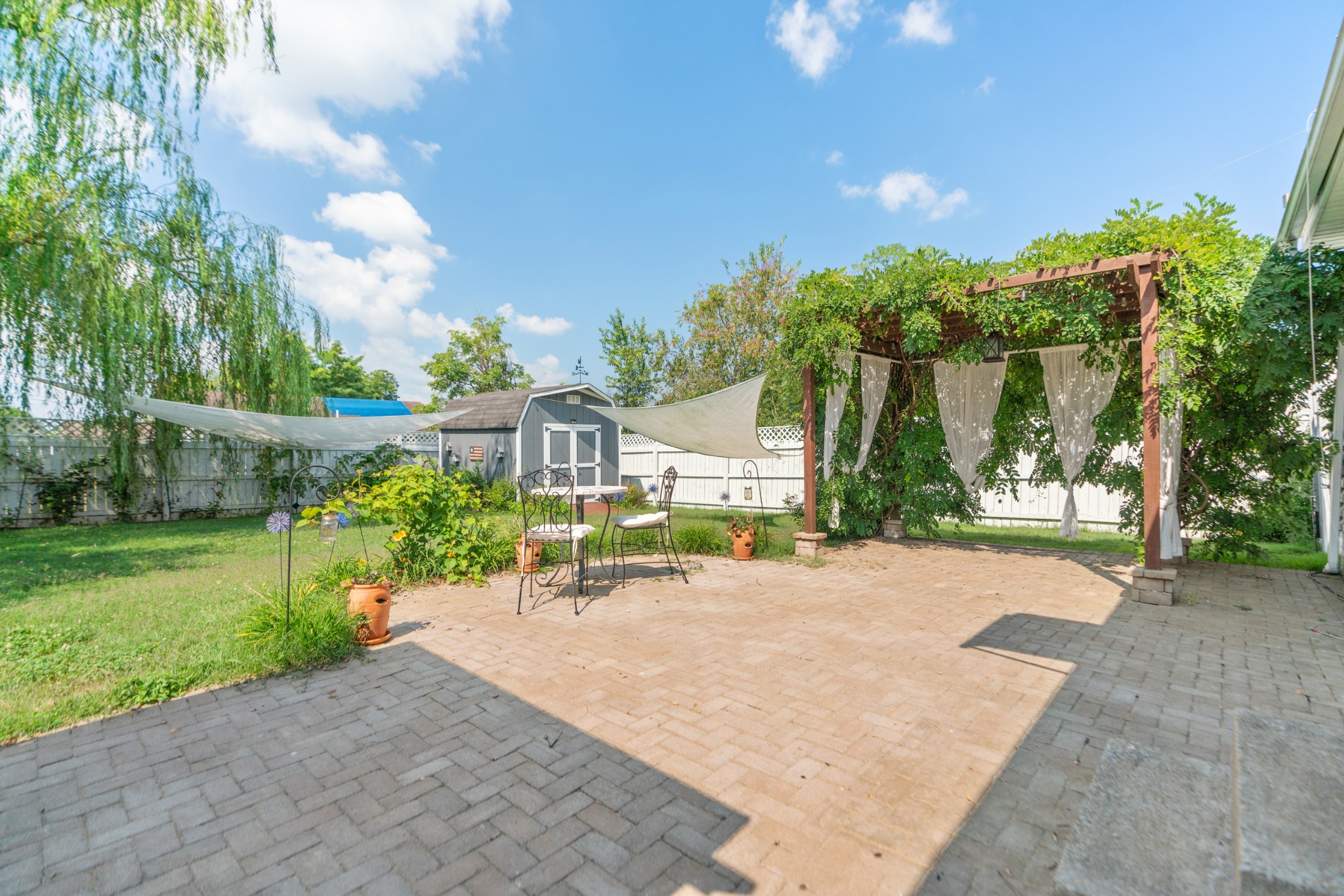 Backyard paver patio with a vine-covered pergola, sheer curtains, and shade sails.