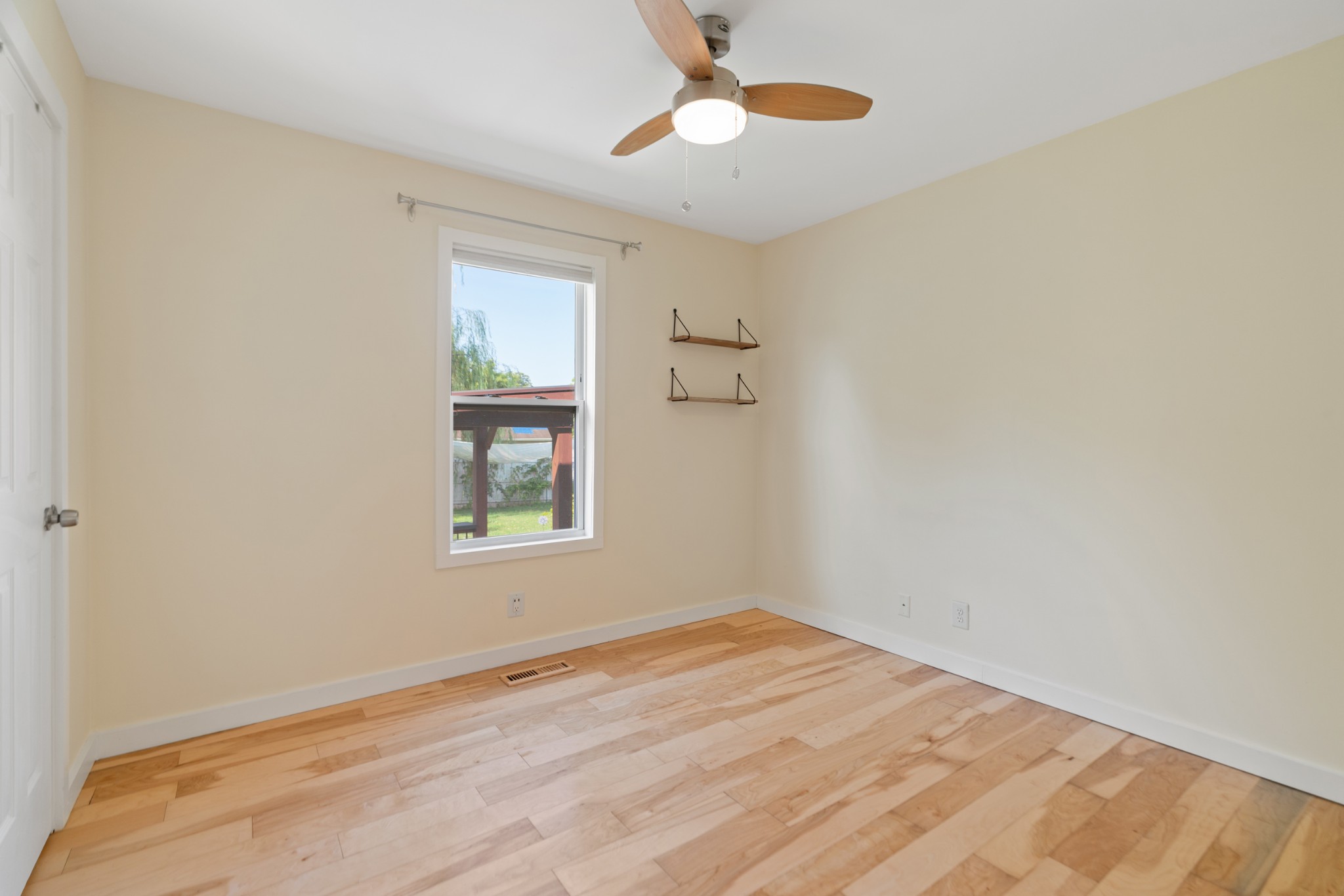 Empty room with light hardwood floors, a ceiling fan, and a window overlooking the backyard.