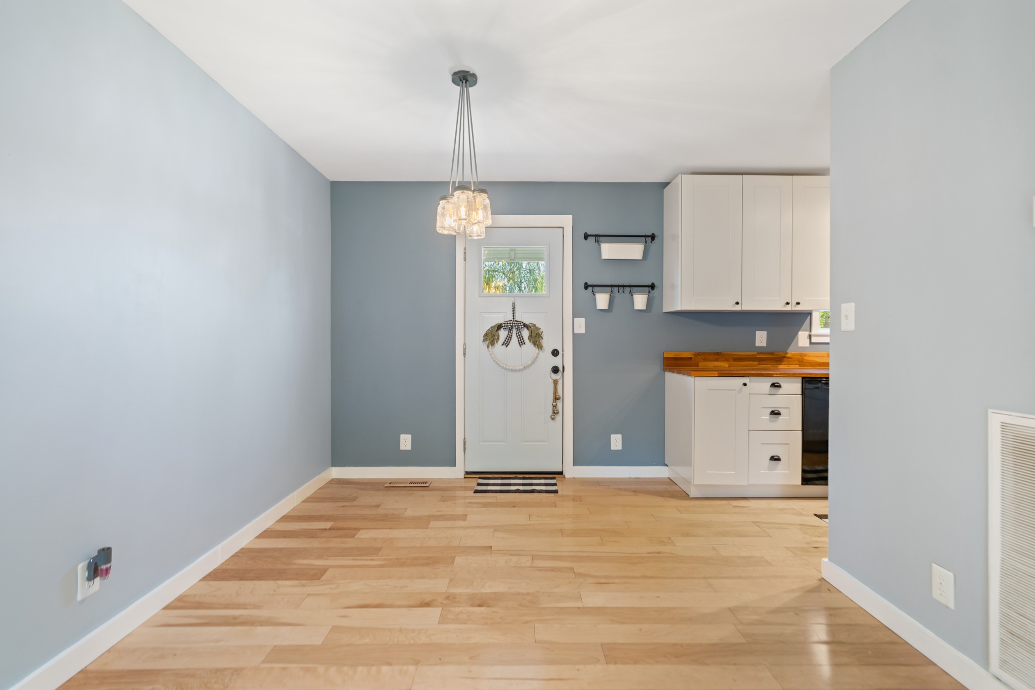 Open dining and entry area with light wood floors, blue walls, a white front door, and adjacent kitchen cabinets.