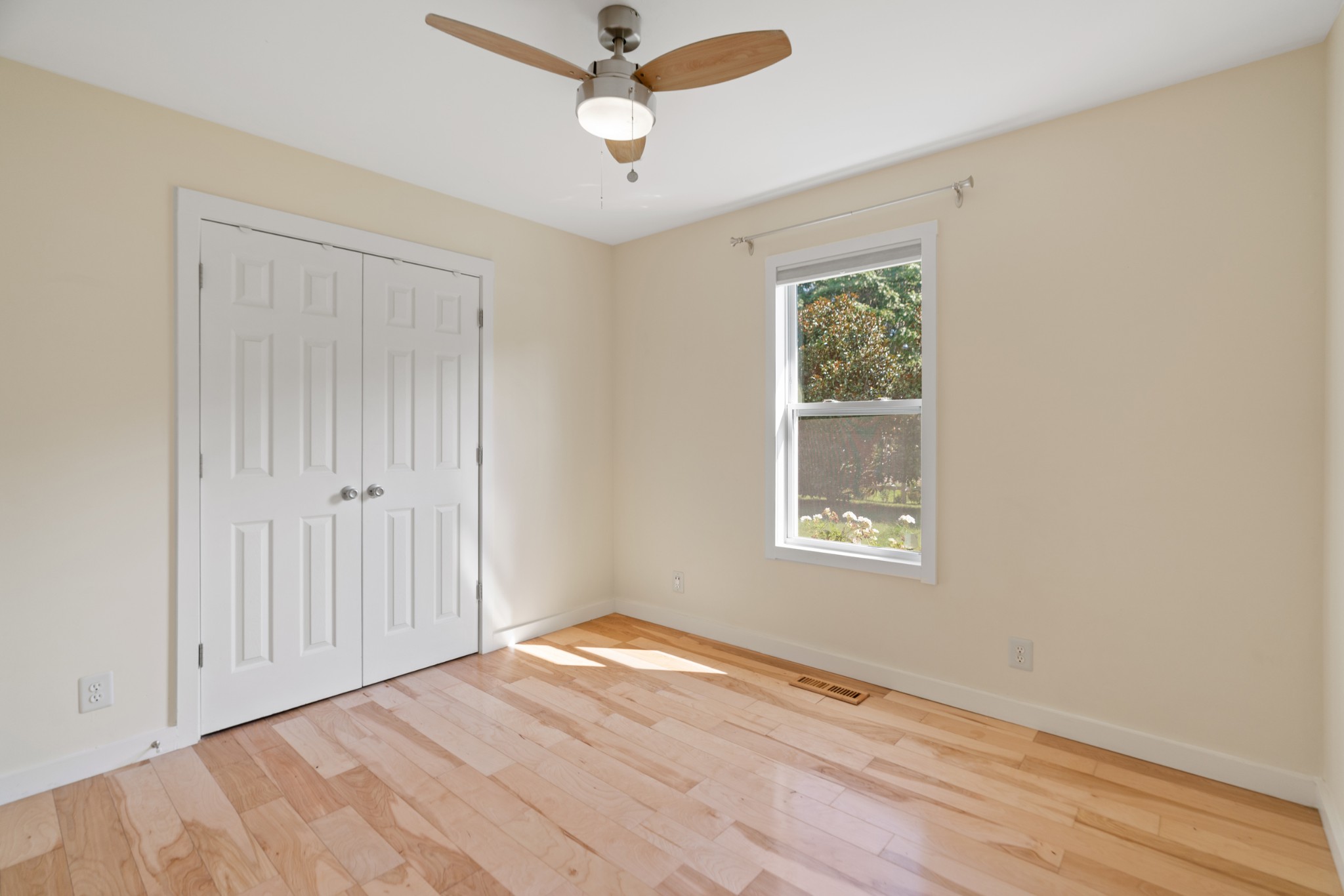 Empty bedroom with hardwood floors, white double closet doors, and a window overlooking greenery.