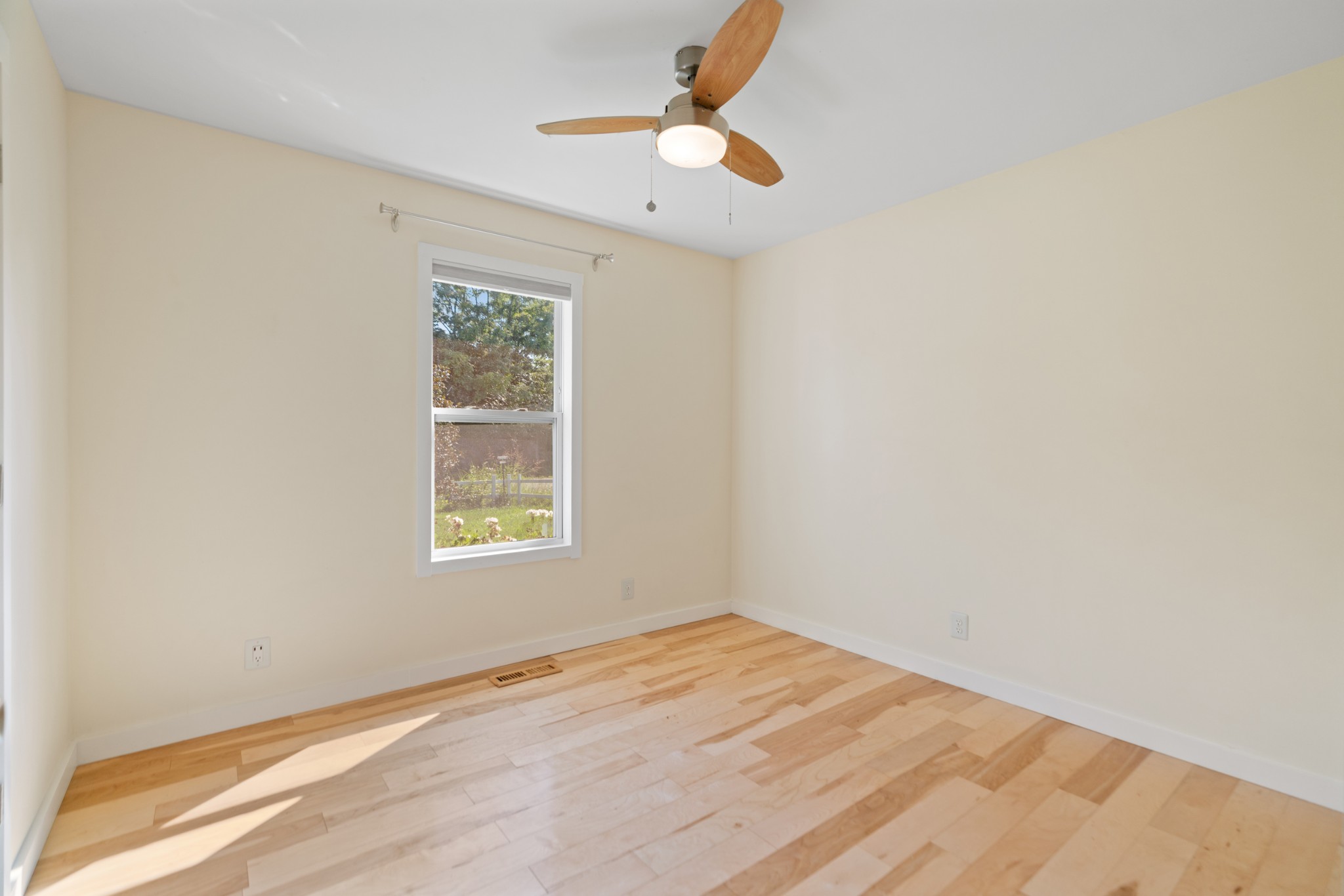 Empty room with light hardwood floors, a window overlooking greenery, and a wooden-blade ceiling fan.