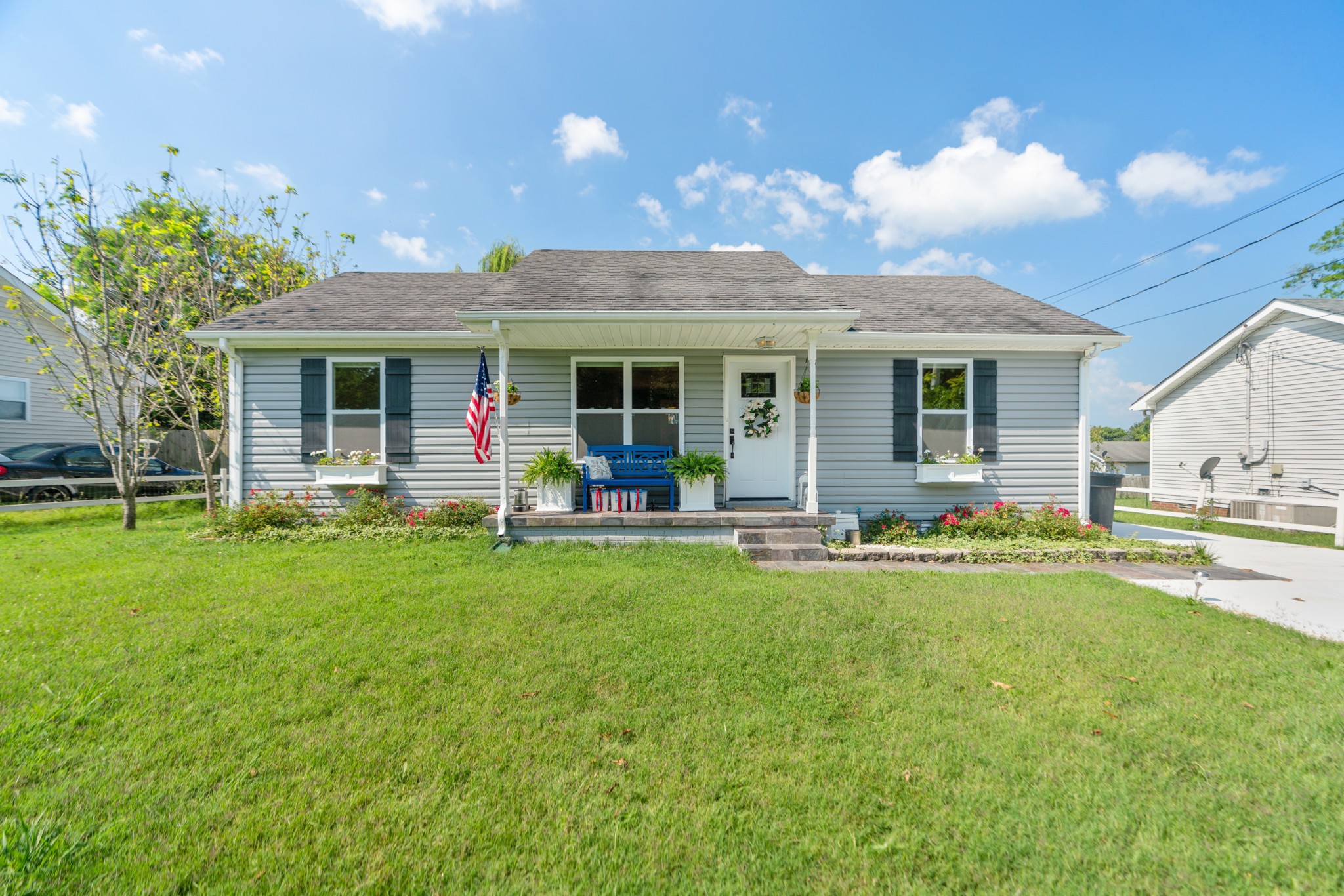 Gray single-story home with a covered porch, blue bench, American flag, and broad green lawn.