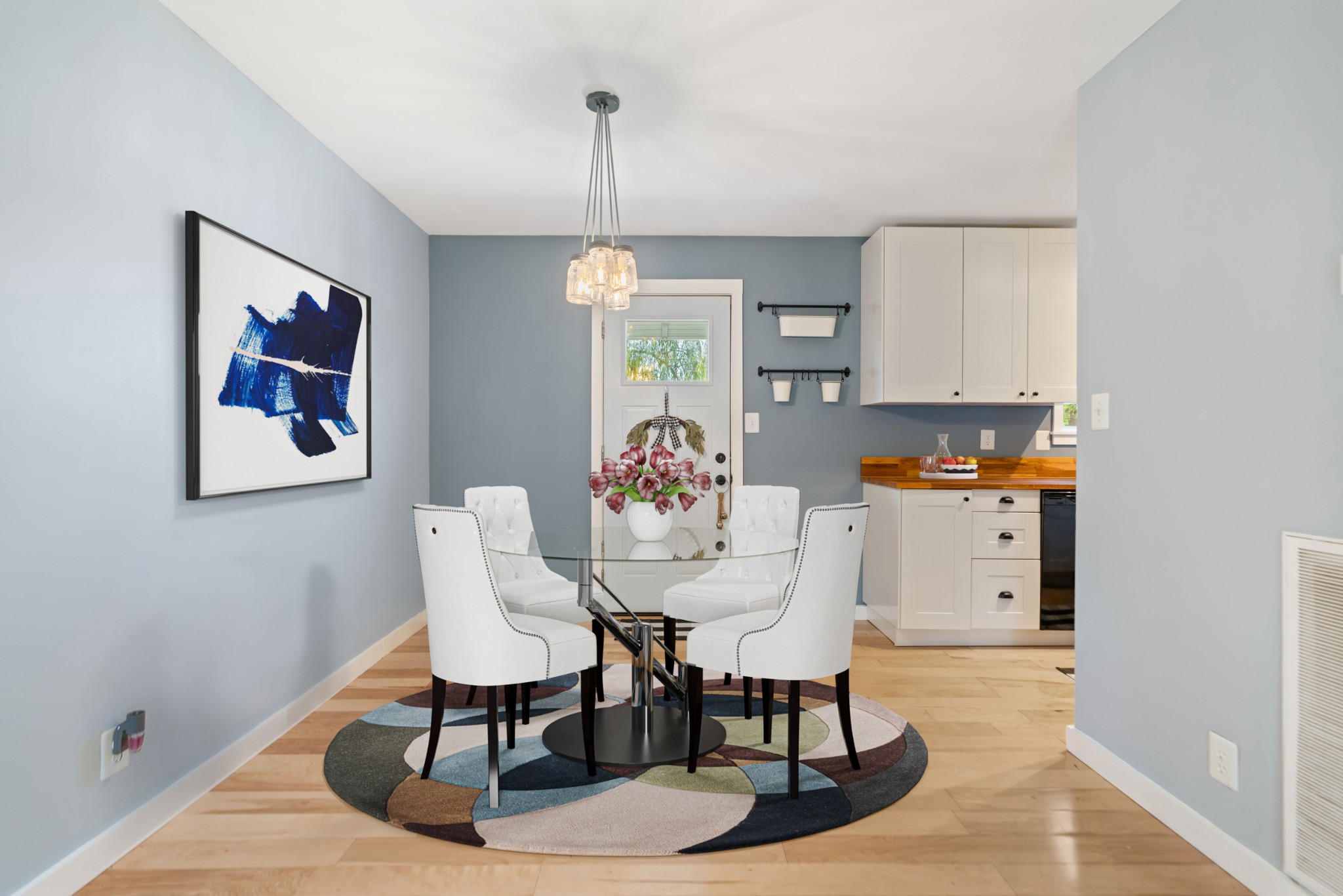 Dining area with a round glass table, white chairs, and a colorful circular rug beside the kitchen.