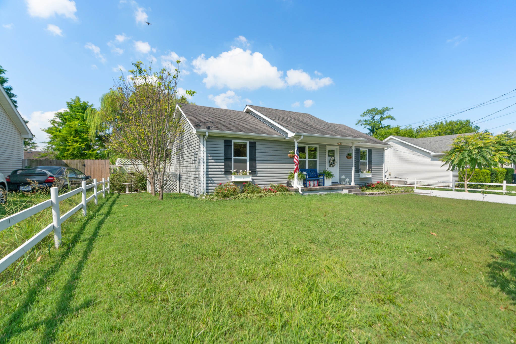 Gray single-story home with a covered porch, blue bench, American flag, and broad front lawn.